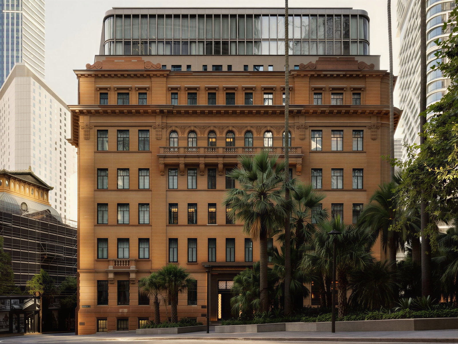 Historic exterior of Capella Sydney with large windows, surrounded by modern skyscrapers and lush palm trees, creating a blend of old and new architecture.