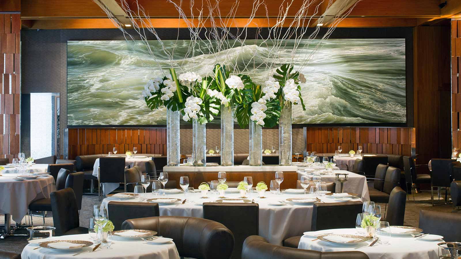 Elegant restaurant with round tables, white orchids in tall vases, and a large ocean wave painting. Sophisticated and serene ambiance.