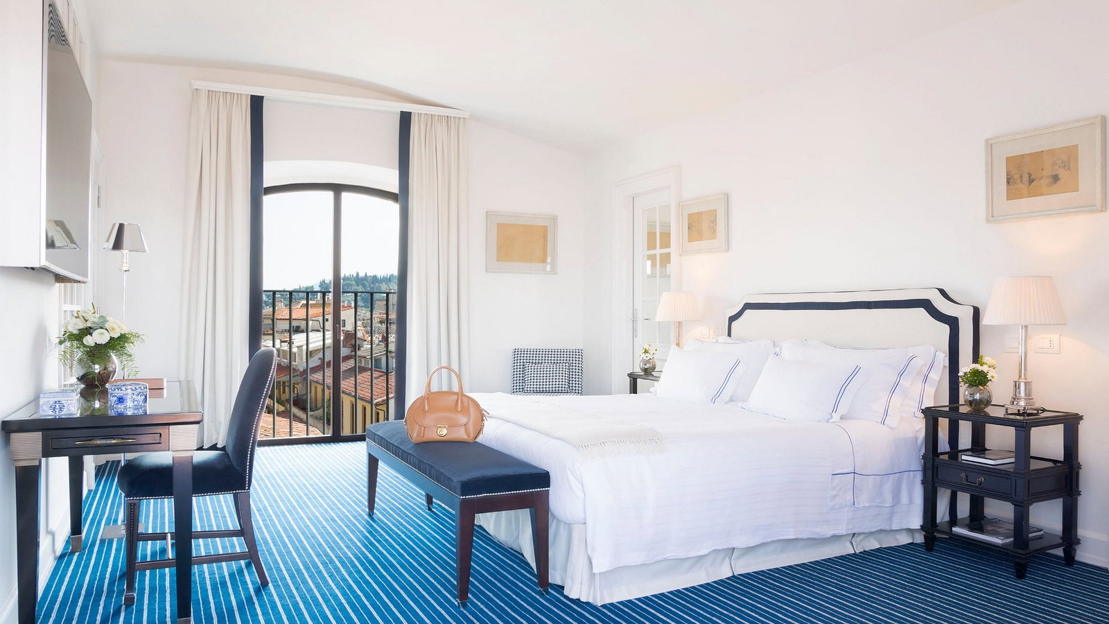 Elegant hotel room with a white bed, blue-striped carpet, and a desk. Large window overlooks rooftops, creating a serene atmosphere.
