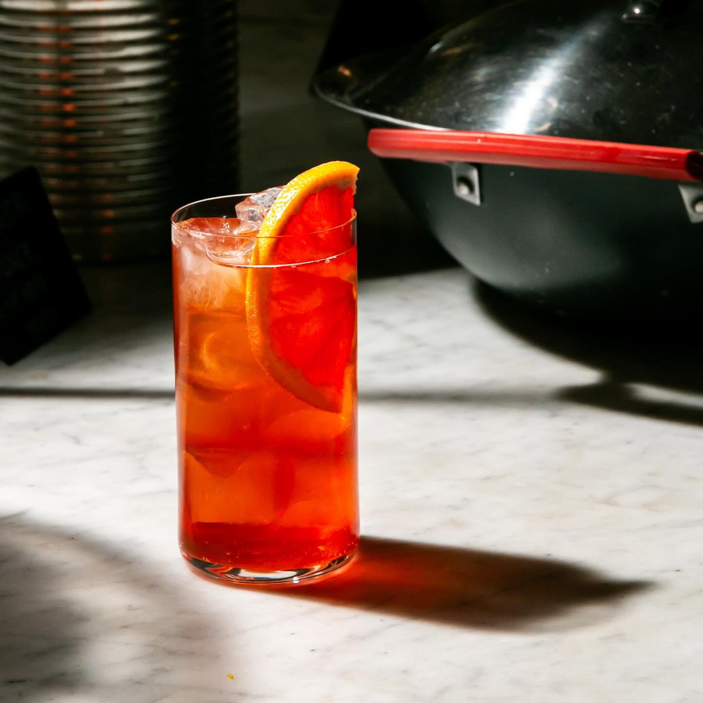 A tall glass of iced red-orange drink with an orange slice, on a marble surface. Warm lighting, cooking pot in the blurred background.