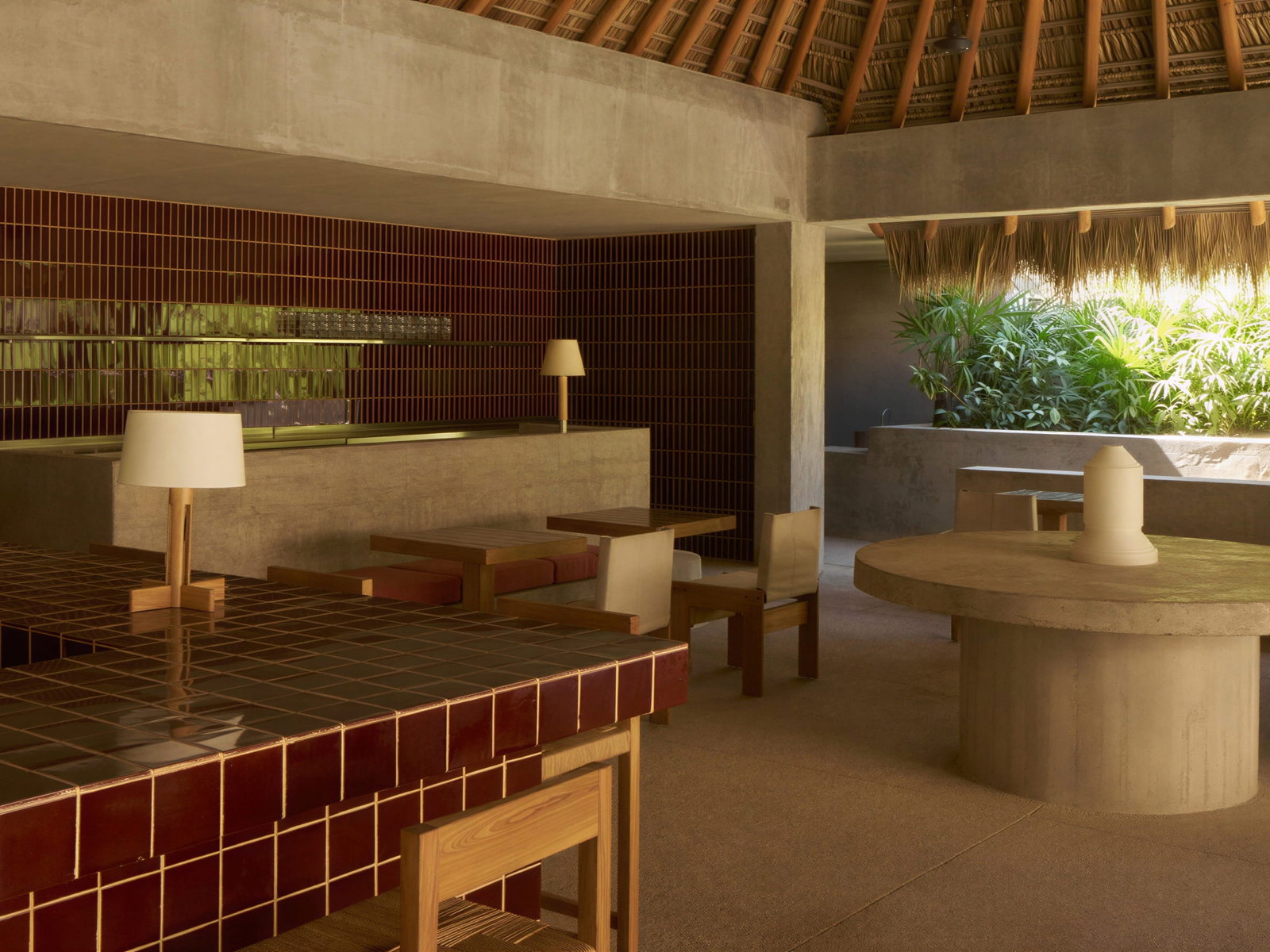 Pool restaurant and bar interior at Hotel Humano with dark red tiles, wooden furniture, and soft lighting. Green plants in sunlight add a refreshing touch.