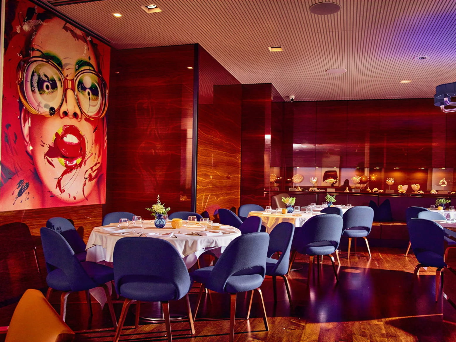Chic interior dining room at Marea NYC with round tables, blue chairs, and vibrant artwork of a woman's face. Warm lighting and minimalist decor.
