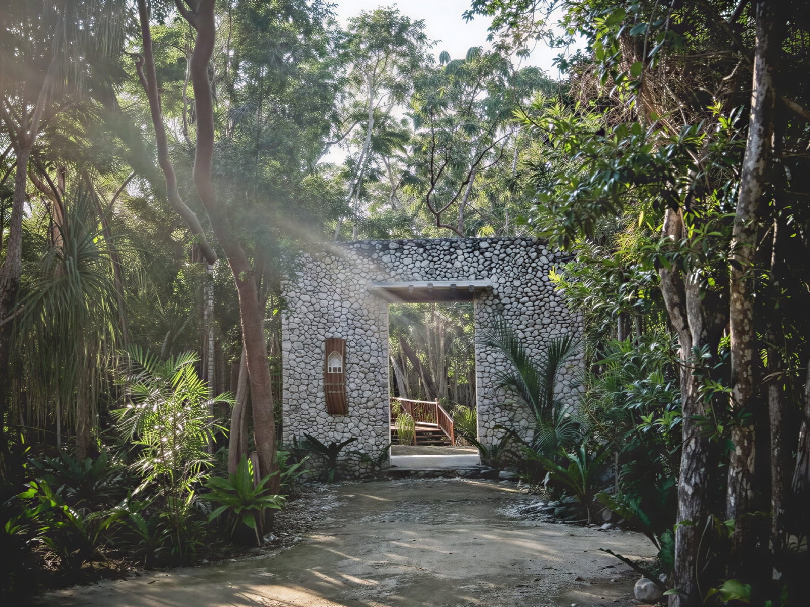 Stone archway at Wakax Hacienda in lush forest, sunlight streaming through trees. Green plants and trees surround the path leading beyond the arch. Serene mood.