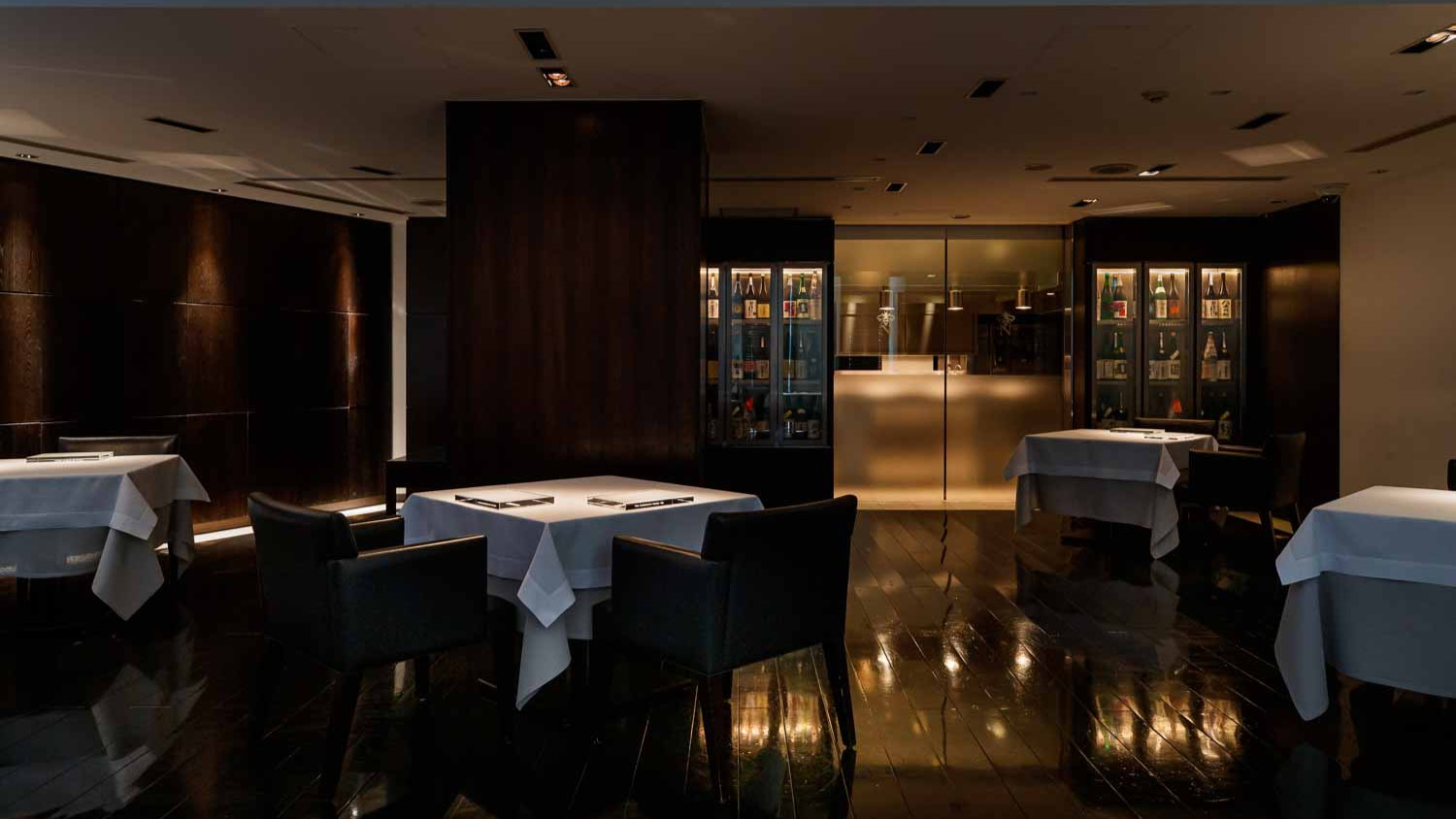 Dimly lit, modern restaurant with dark wood accents, white tablecloths on tables, and display cabinets with bottles. Mood is elegant and calm.