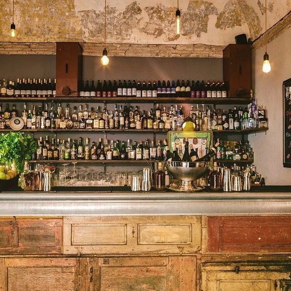 Rustic bar with a weathered counter, shelves stocked with liquor bottles, hanging lights, chalkboard menu, and a basket of green apples.