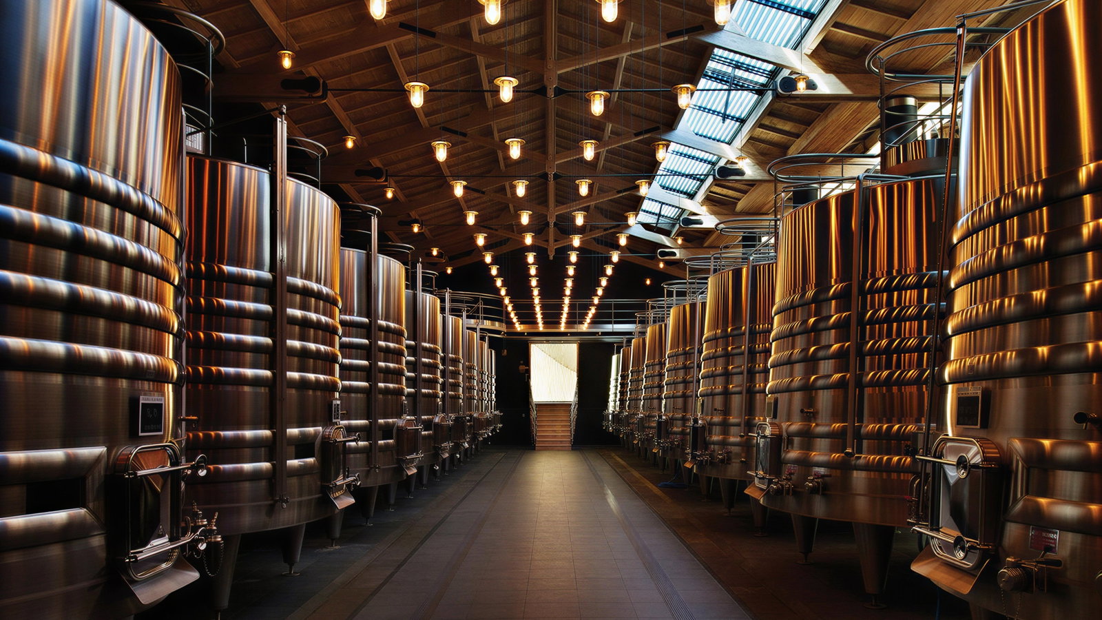 Large, shiny metal fermentation tanks line both sides of an illuminated winery interior with wooden ceiling and skylights. Calm, industrial mood.