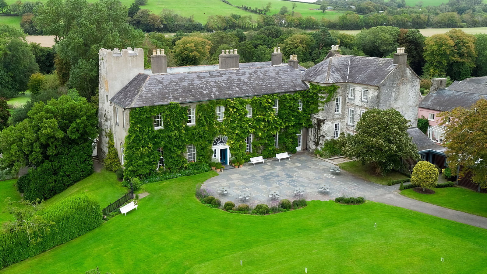 Historic mansion with ivy-clad walls, set in lush gardens. Stone courtyard with benches and tables. Vibrant green landscape surrounds.