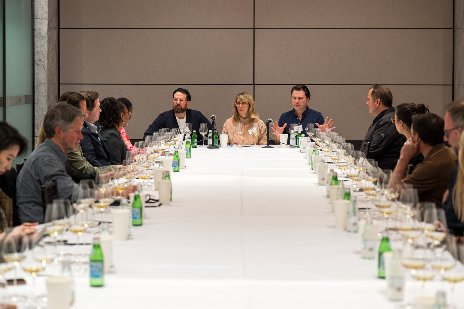 Panel discussion at a long table with three speakers; attendees listen attentively. Bottled water and wine glasses are neatly arranged.