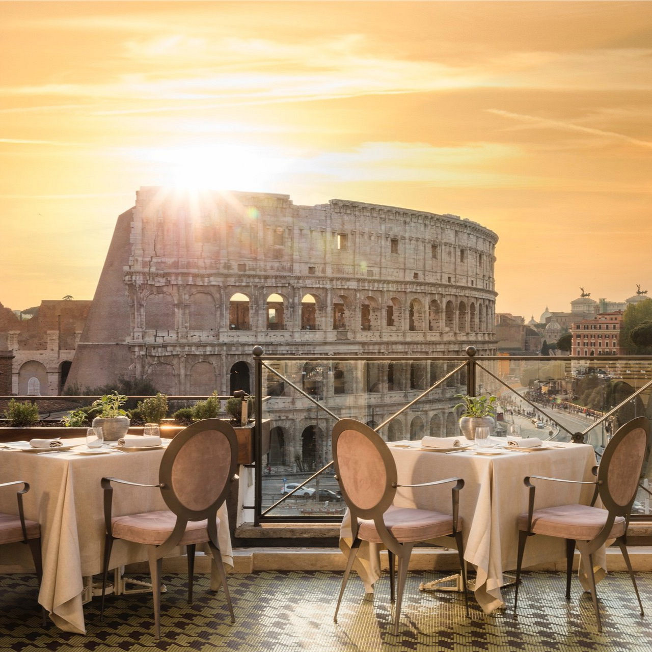 Sunset view from a terrace overlooking the Colosseum. Elegant tables with white cloths and soft chairs create a serene ambiance.