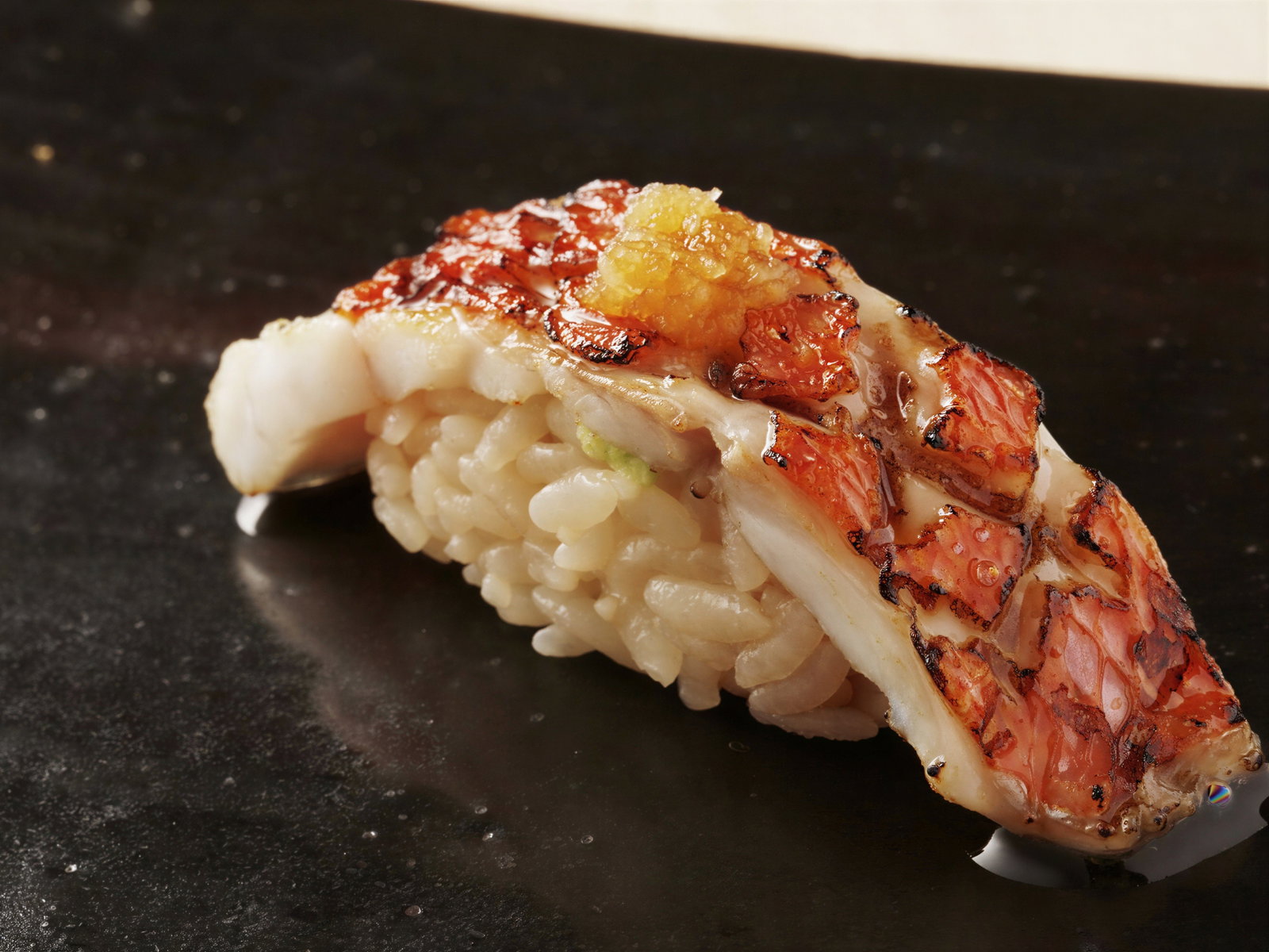 Grilled sushi piece with red fish atop white rice on a glossy black plate, garnished with golden sauce, creating a refined and elegant look.