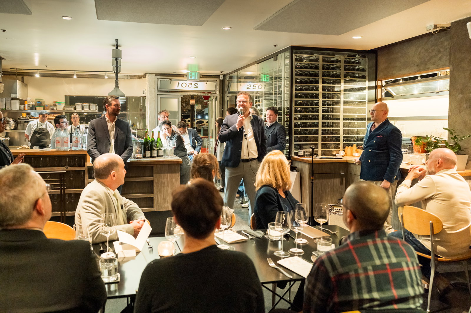 A speaker addresses a seated group in a restaurant. Men in suits stand by; kitchen staff are in the background. Wine glasses and tables visible.