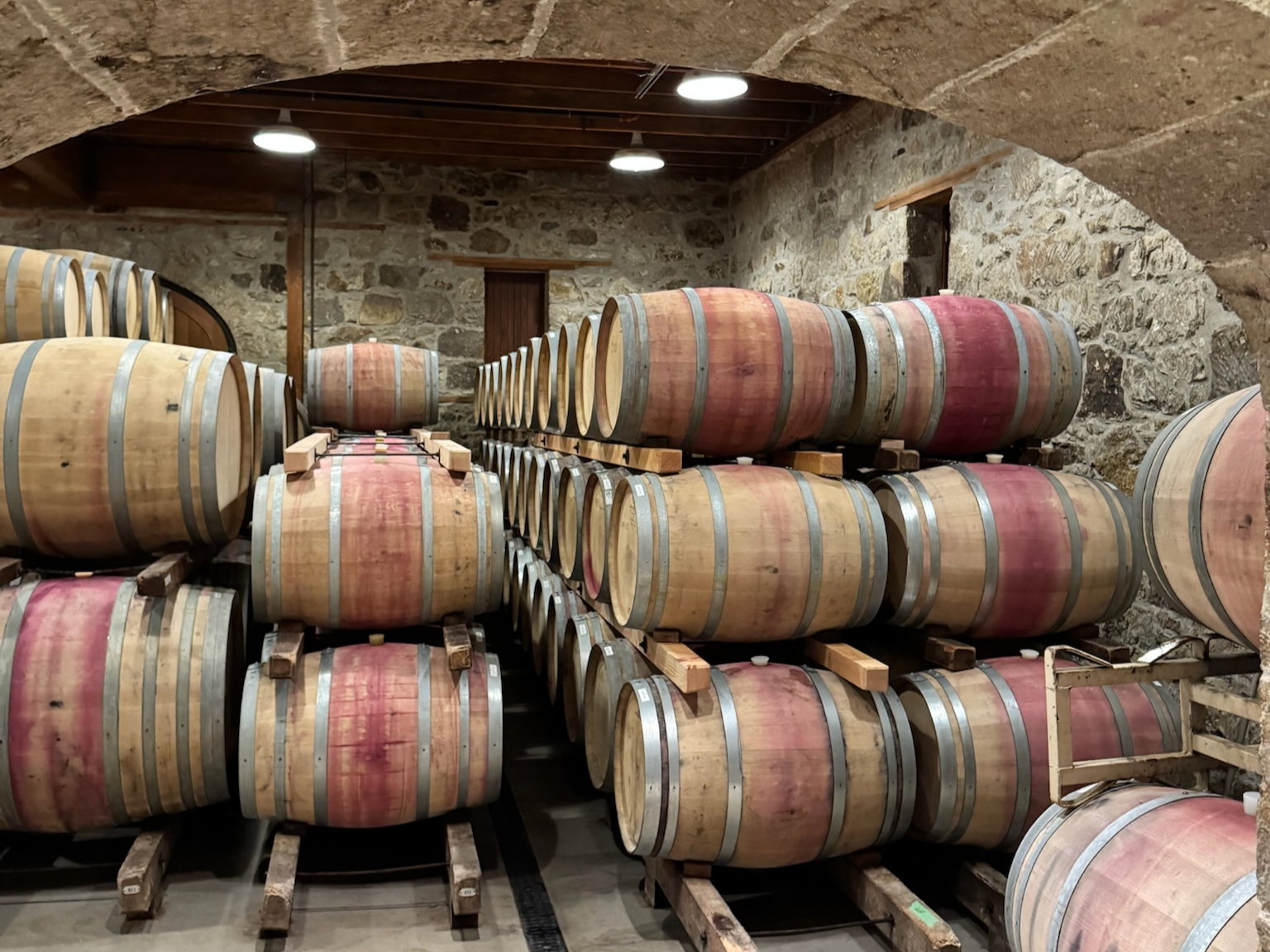 Wooden wine barrels stacked in a stone cellar. The barrels have red stains, creating a rustic, vintage atmosphere. No text visible.