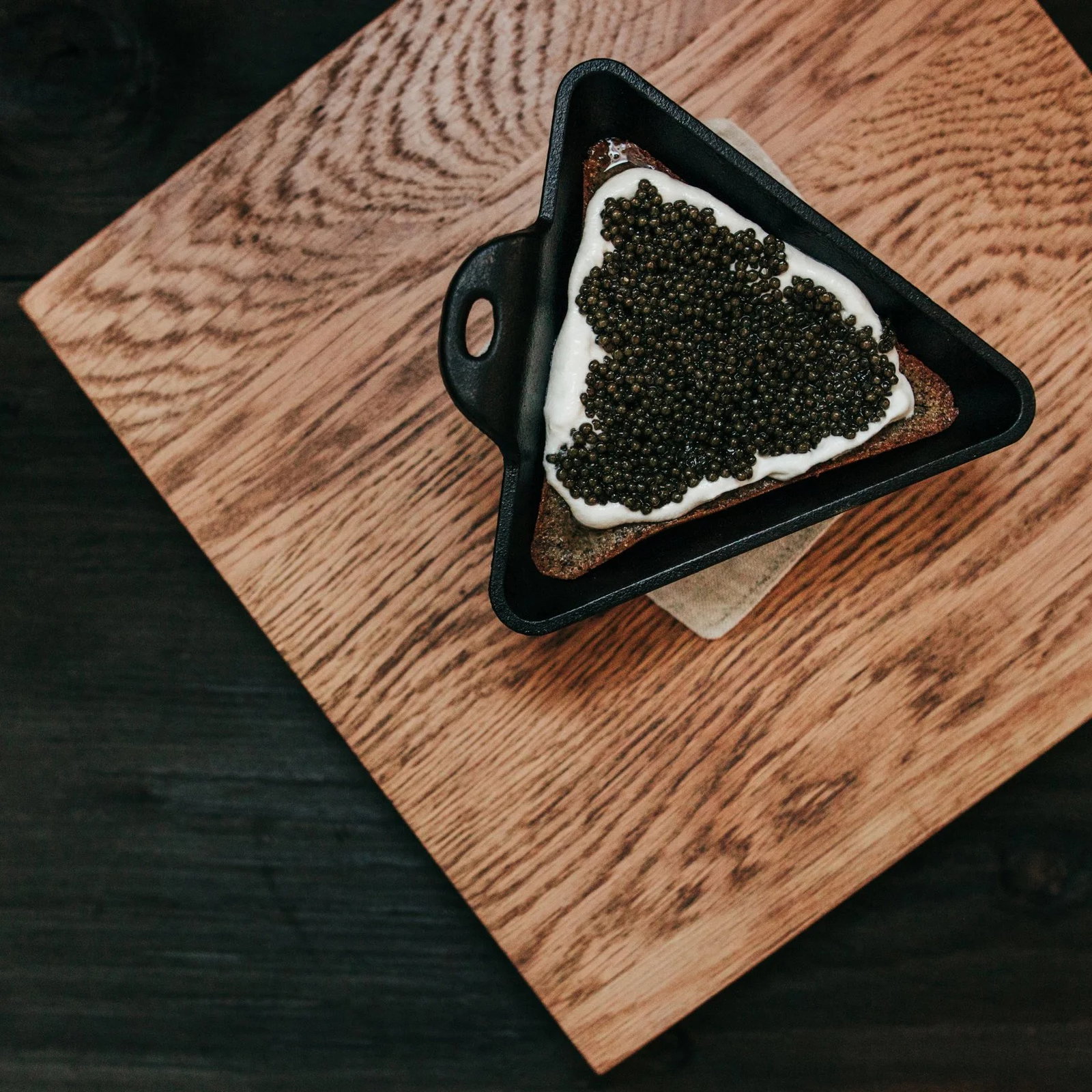 A triangle toast with caviar and cream in a black dish sits on a textured wooden board, showcasing a luxurious culinary presentation.