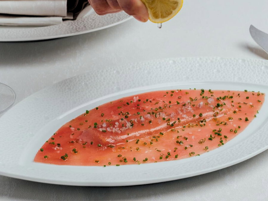Sliced tuna with herbs on a white plate; hand squeezes lemon juice over it. Soft lighting, elegant table setting.