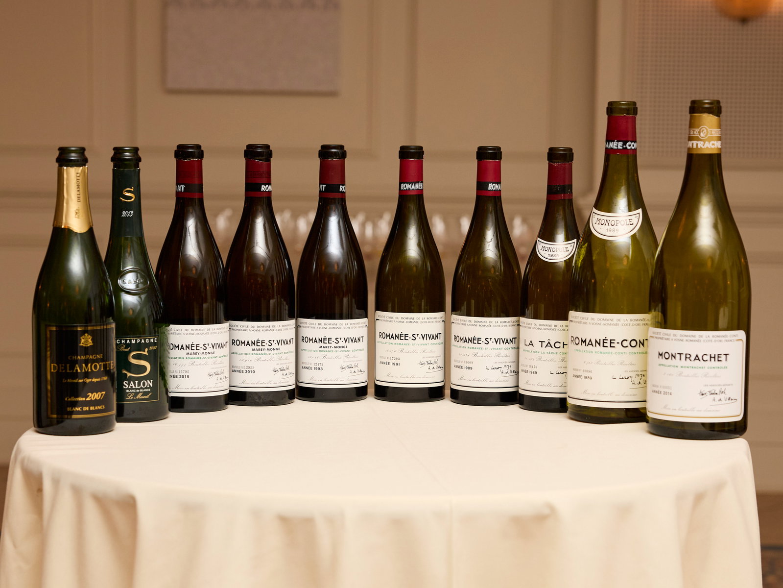 Ten wine bottle lineup from The Legends Dinner at La Paulée NYC 2025 with labels including Romanée-Conti and Montrachet are arranged on a round table with a white tablecloth in a softly lit room.