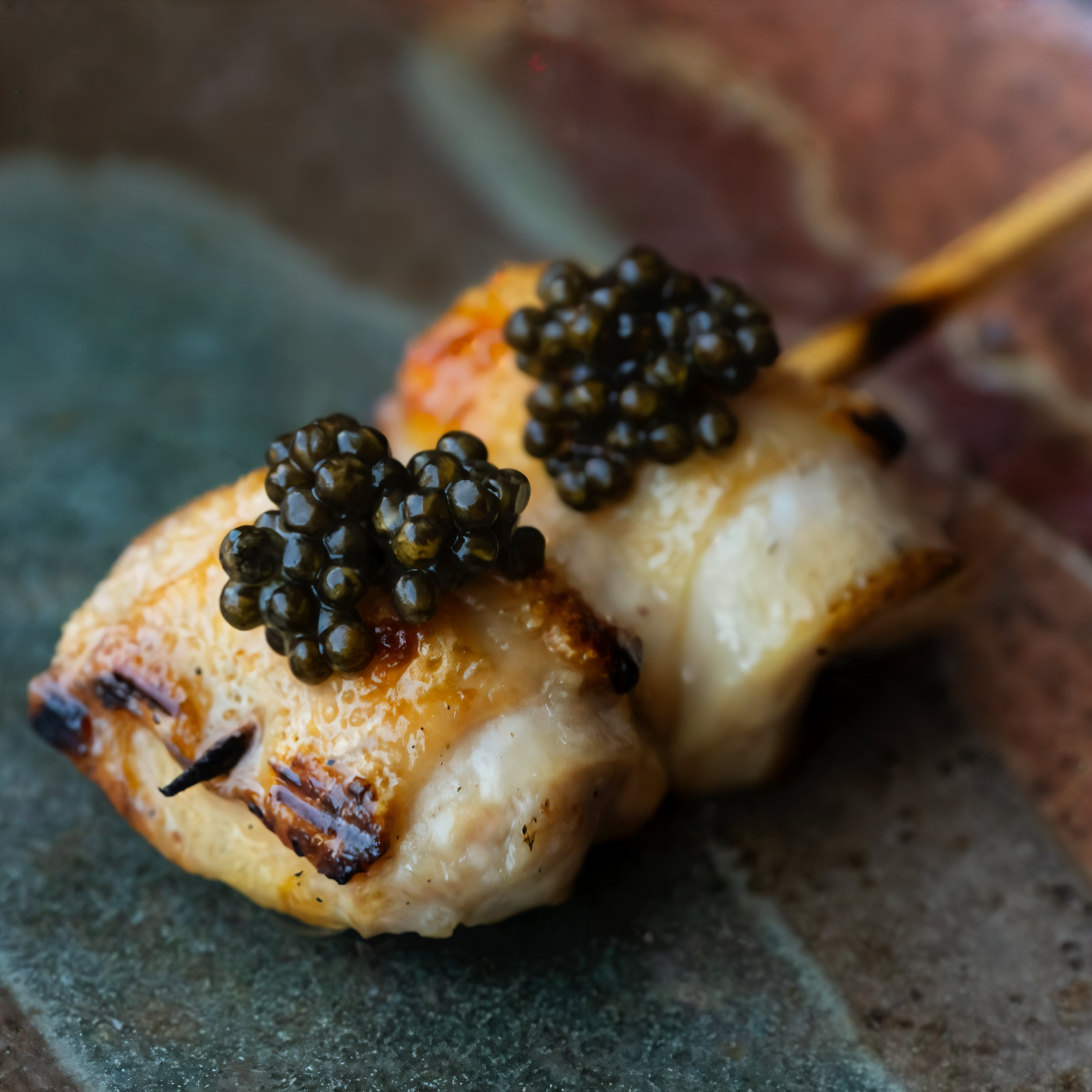 Grilled skewered chicken topped with black caviar on a rustic ceramic plate, featuring golden brown edges and a speckled surface.