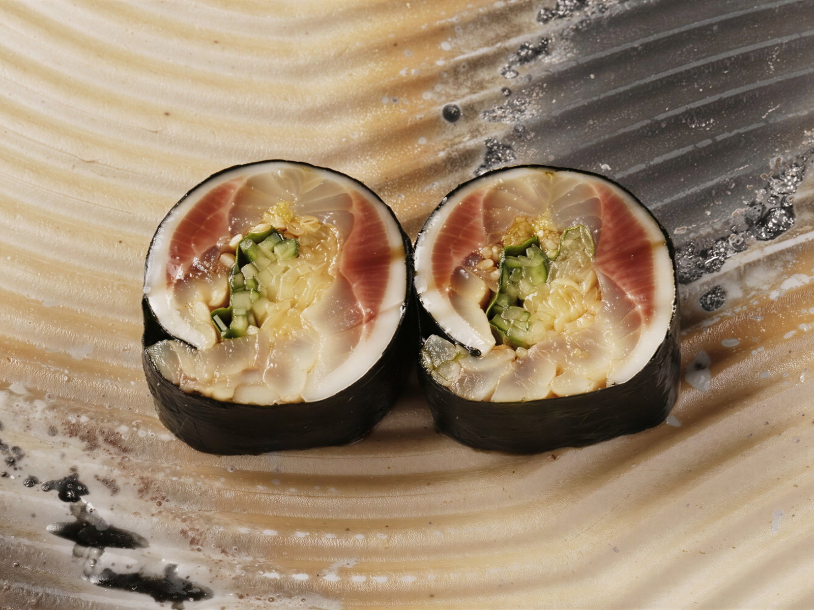 Two sushi rolls with fish, cucumber, and wasabi on a textured, wavy beige and gray plate. The sushi is neatly cut, showcasing vibrant colors.