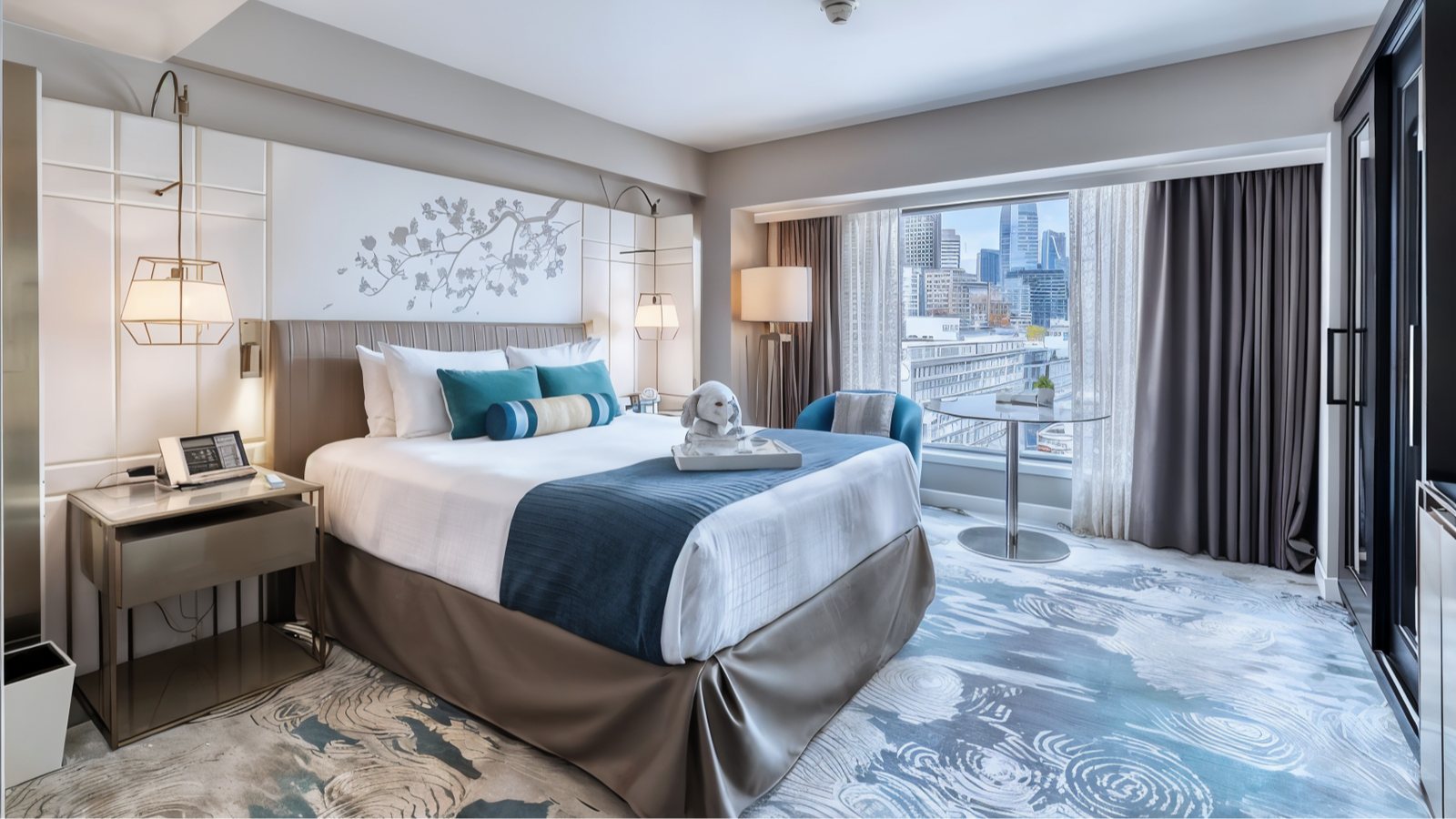 Stylish hotel room with a bed, blue accents, plush toys, modern decor, and city view through large windows. Calm, inviting atmosphere.