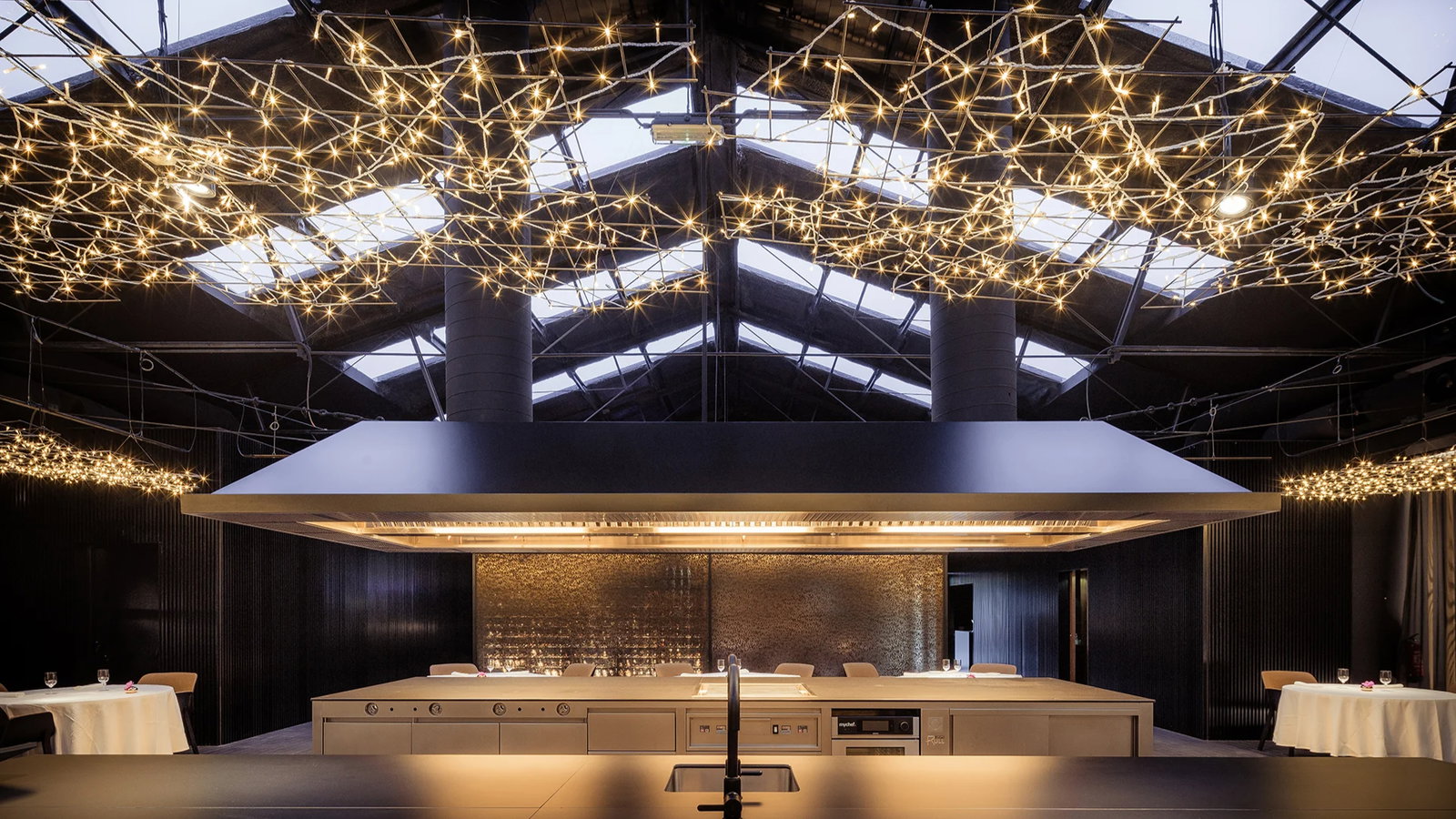 Modern restaurant interior with star-like ceiling lights, large central kitchen counter, and elegant table settings. Dark, sophisticated ambiance.