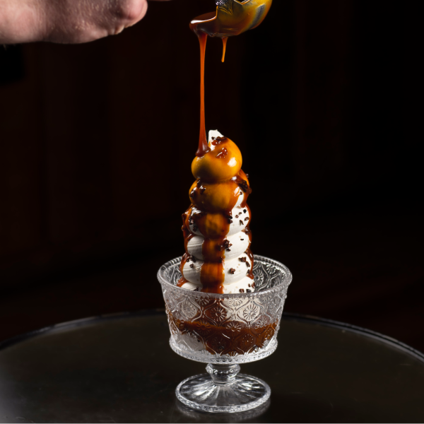 Caramel sauce is drizzled over a tall swirl of ice cream in an ornate glass dish, creating a rich, indulgent dessert against a dark background.