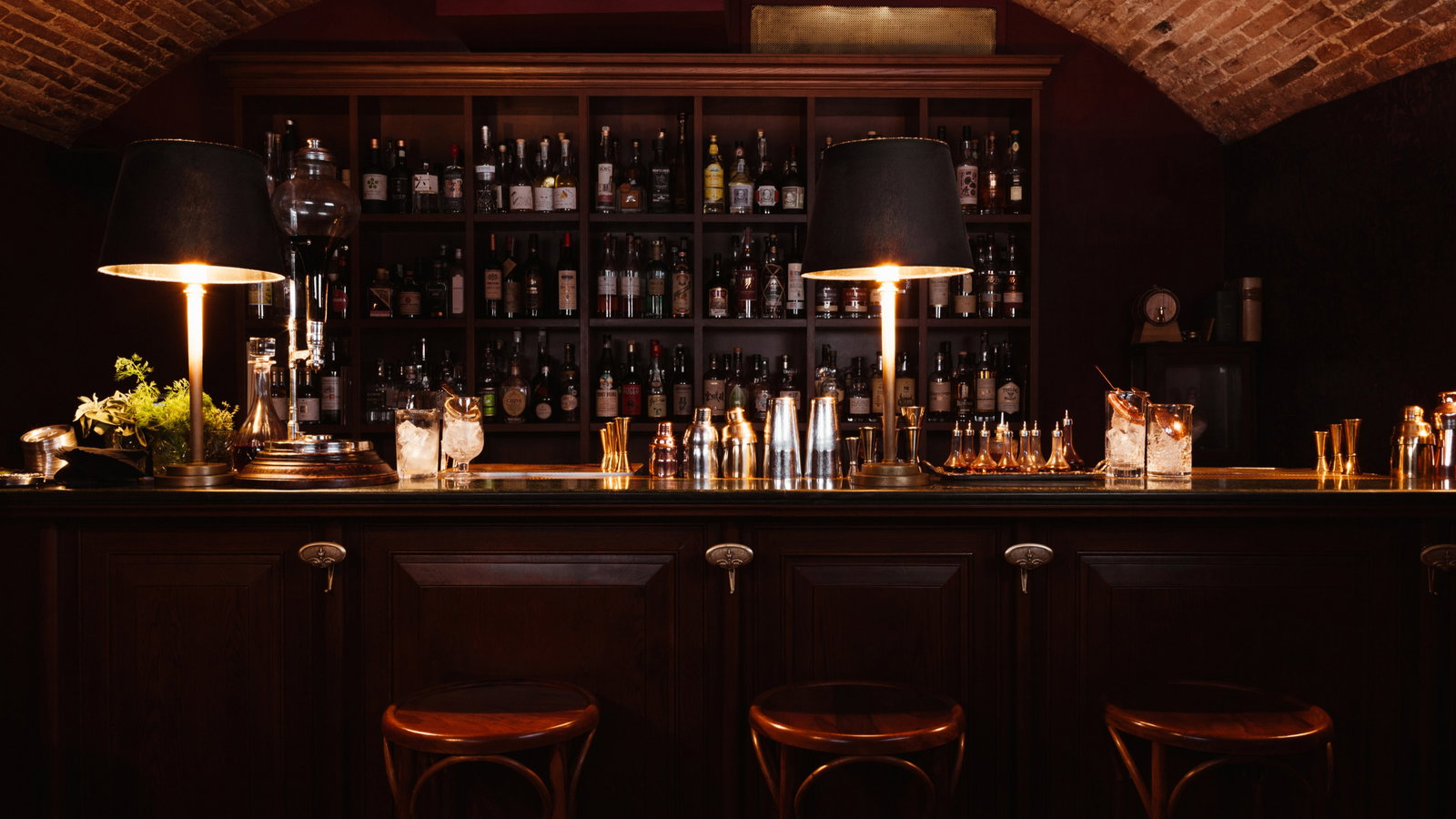 Dimly lit bar with dark wood, shelves of bottles, two lamps, and copper bar tools. Two ice-filled glasses sit on the counter. Cozy ambiance.