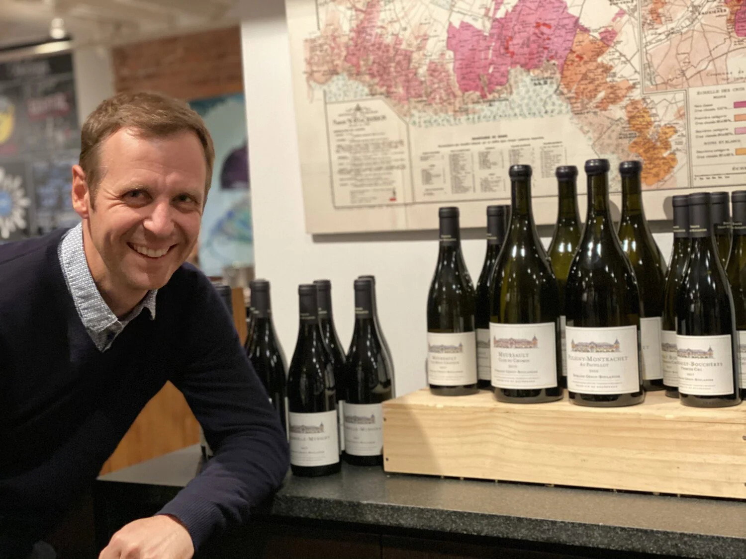 Domaine Génot-Boulanger winemaker Guillaume Lavolée smiles next to wine bottles on a counter, with a colorful map in the background. The setting is warm and inviting.