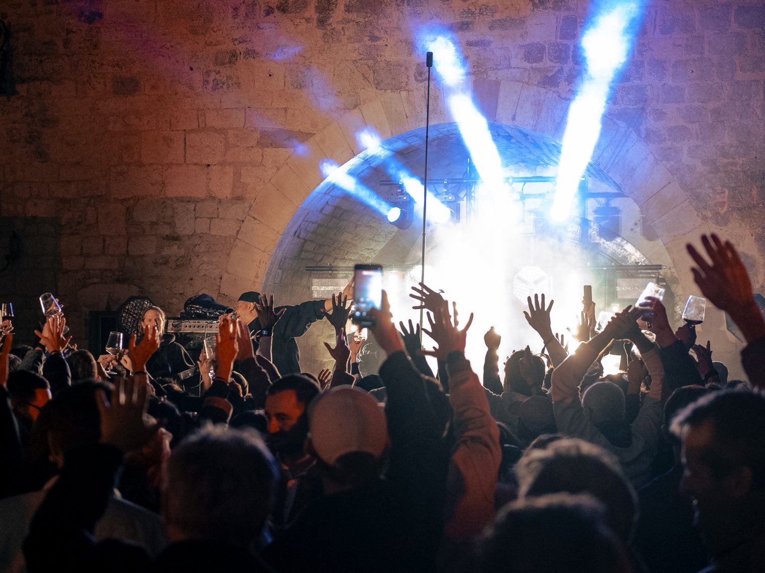 Vin et hip hop (wine and hip hop) Concert crowd in burgundy raises hands and phones in front of stone wall. Bright stage lights create beams. Energetic atmosphere.