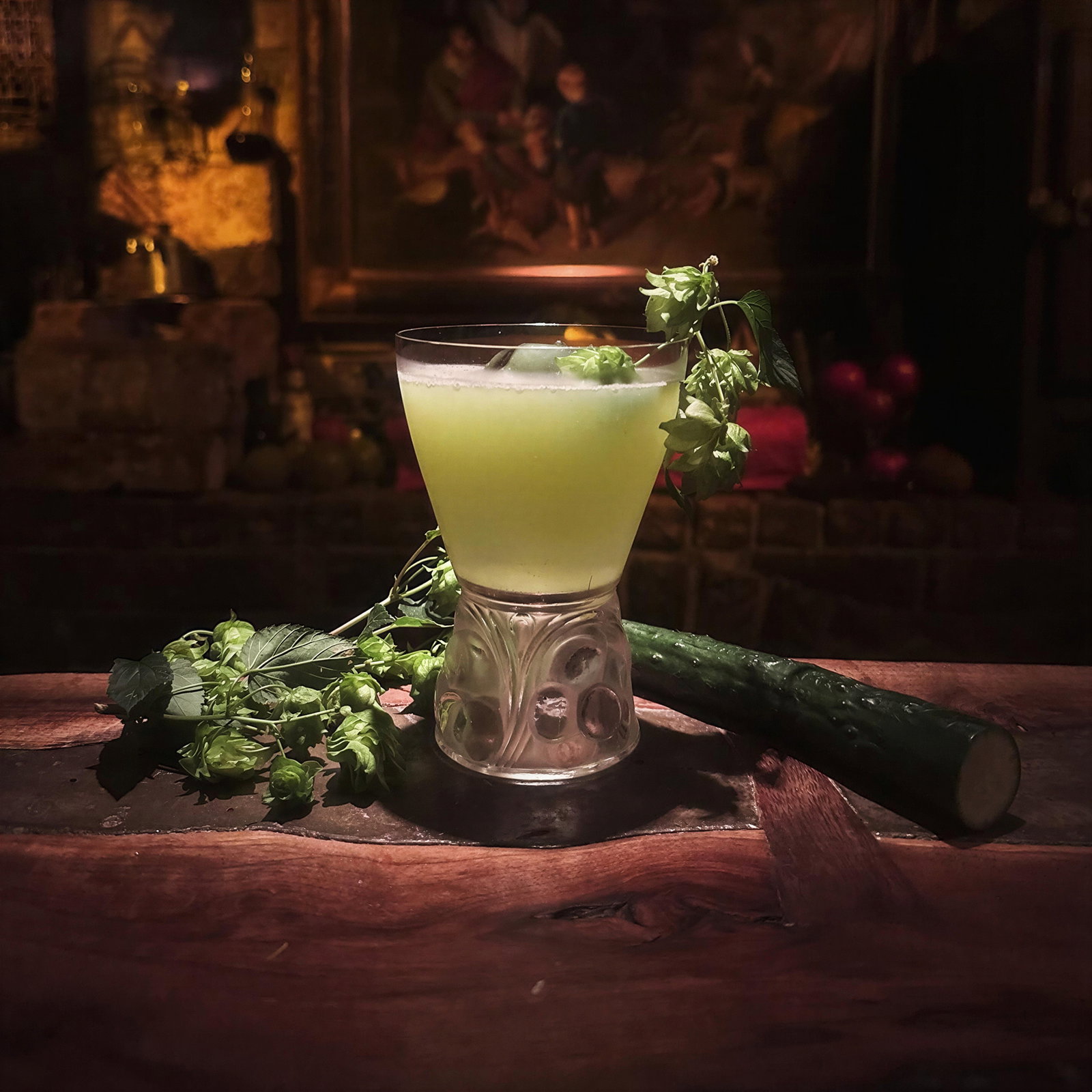 A green drink garnished with herbs sits in a decorative glass on a wooden table. A cucumber and foliage are nearby; a painting is blurred in the background.
