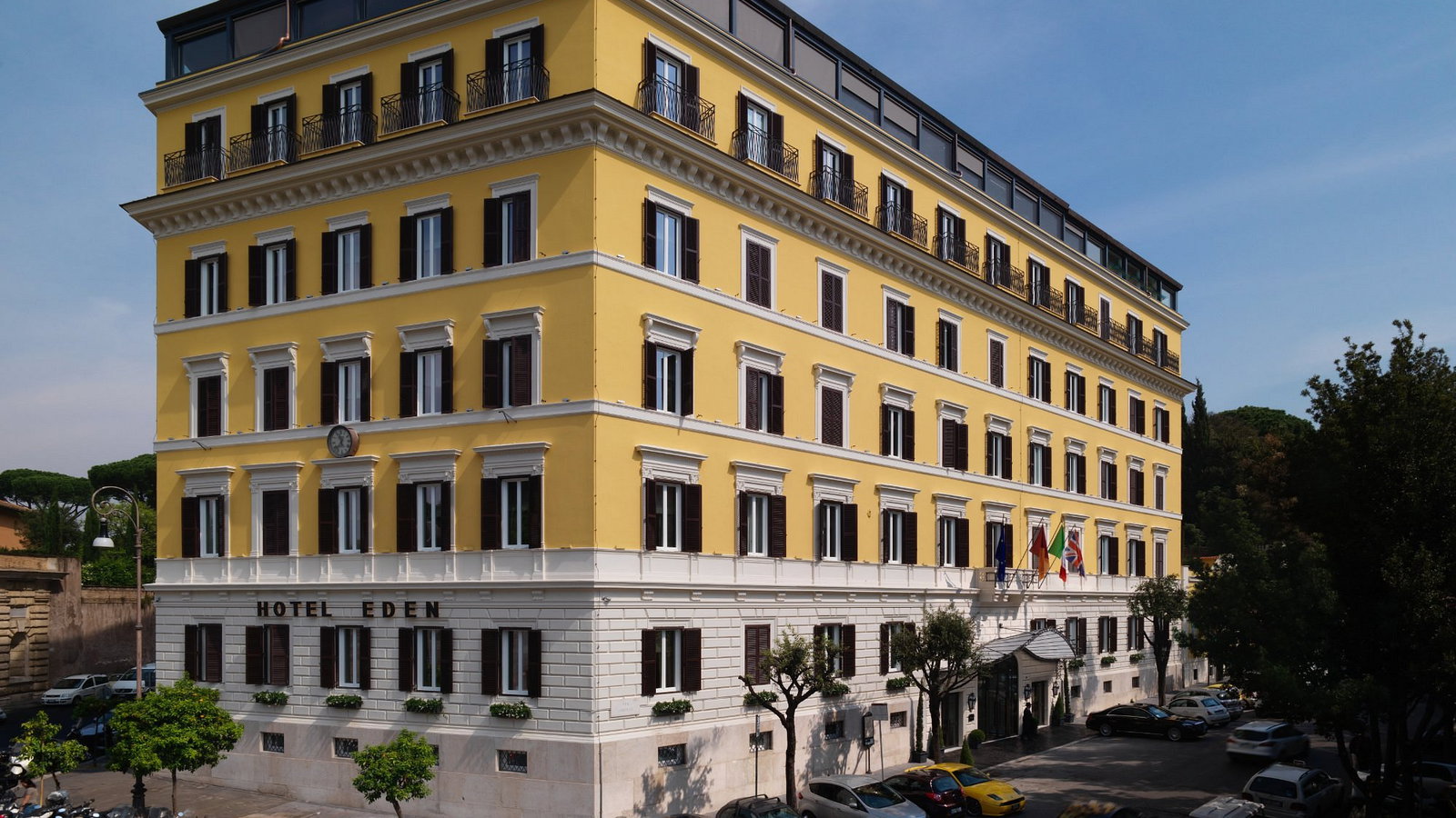 Yellow hotel building with black shutters, labeled "Hotel Eden." Flags are displayed, trees and parked cars line the street below.