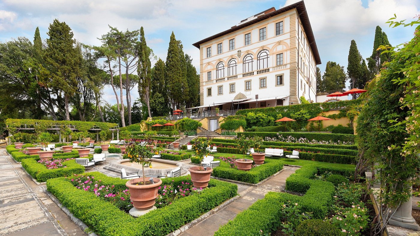 Elegant villa with lush gardens, terracotta pots, and vibrant flowers. Red umbrellas dot the green landscape under a partly cloudy sky.