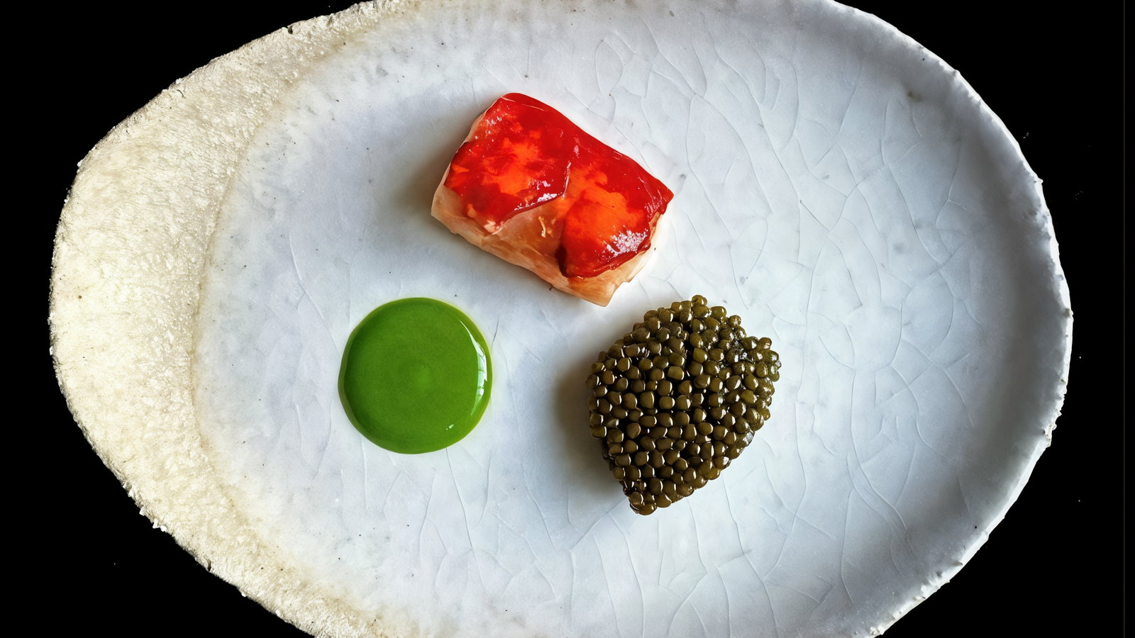 Gourmet dish with red-topped fish, green sauce, and caviar on a textured white plate against a black background. Elegant presentation.