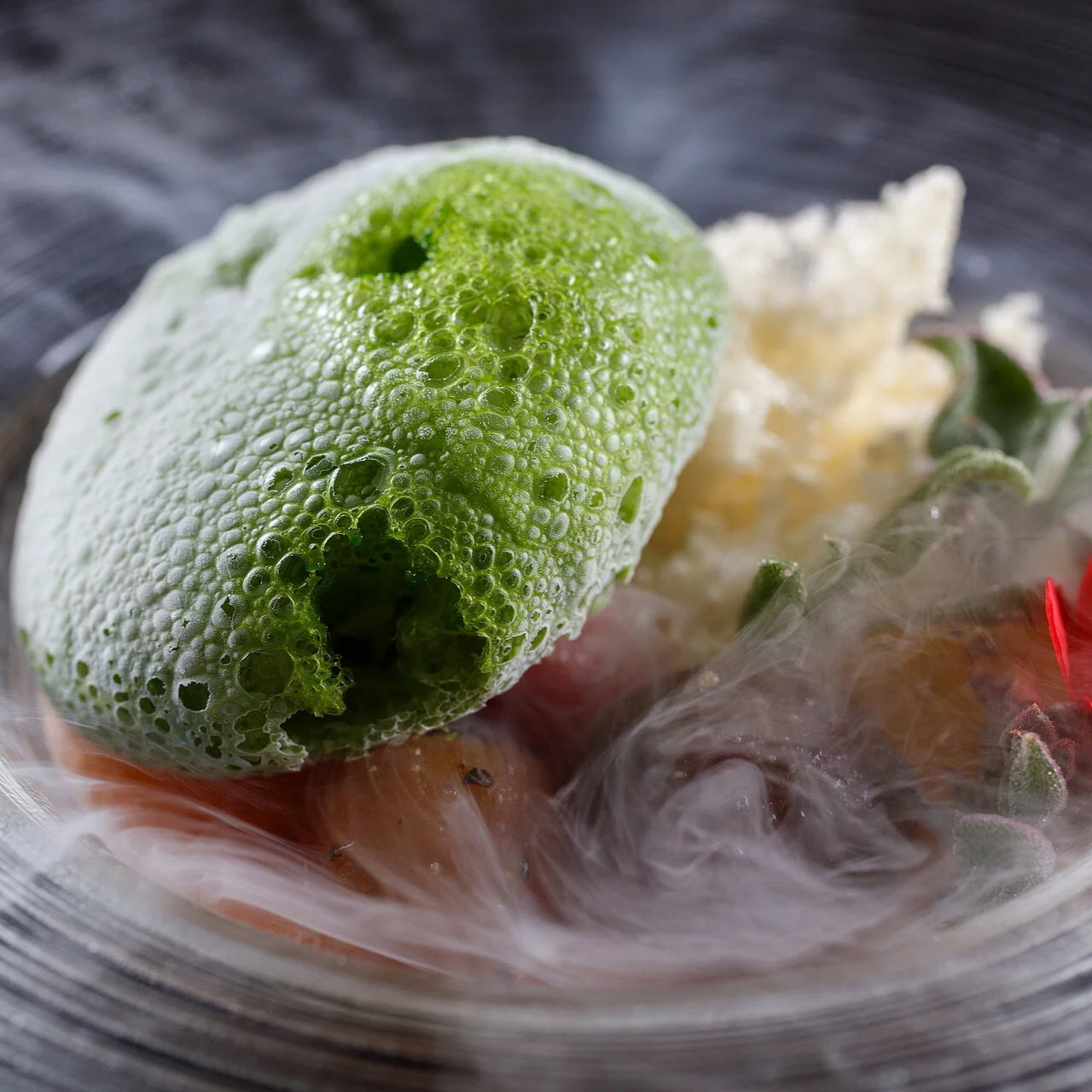 Green foam atop food garnished with herbs, surrounded by mist, on a grey textured plate. The scene is artistic and atmospheric.