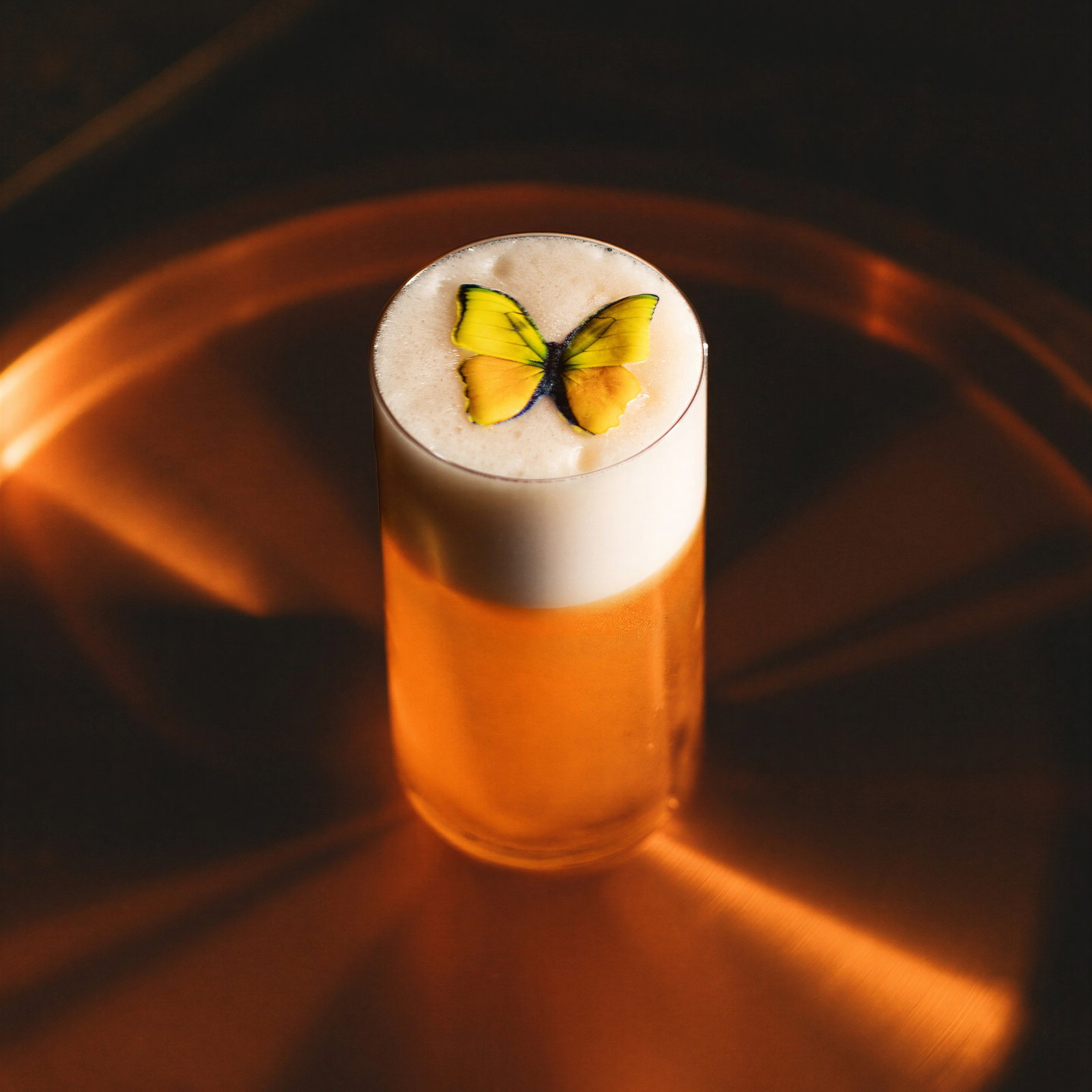 A tall glass of foamy beer with a yellow butterfly decoration on top, set against a warm, diffused copper-toned background.