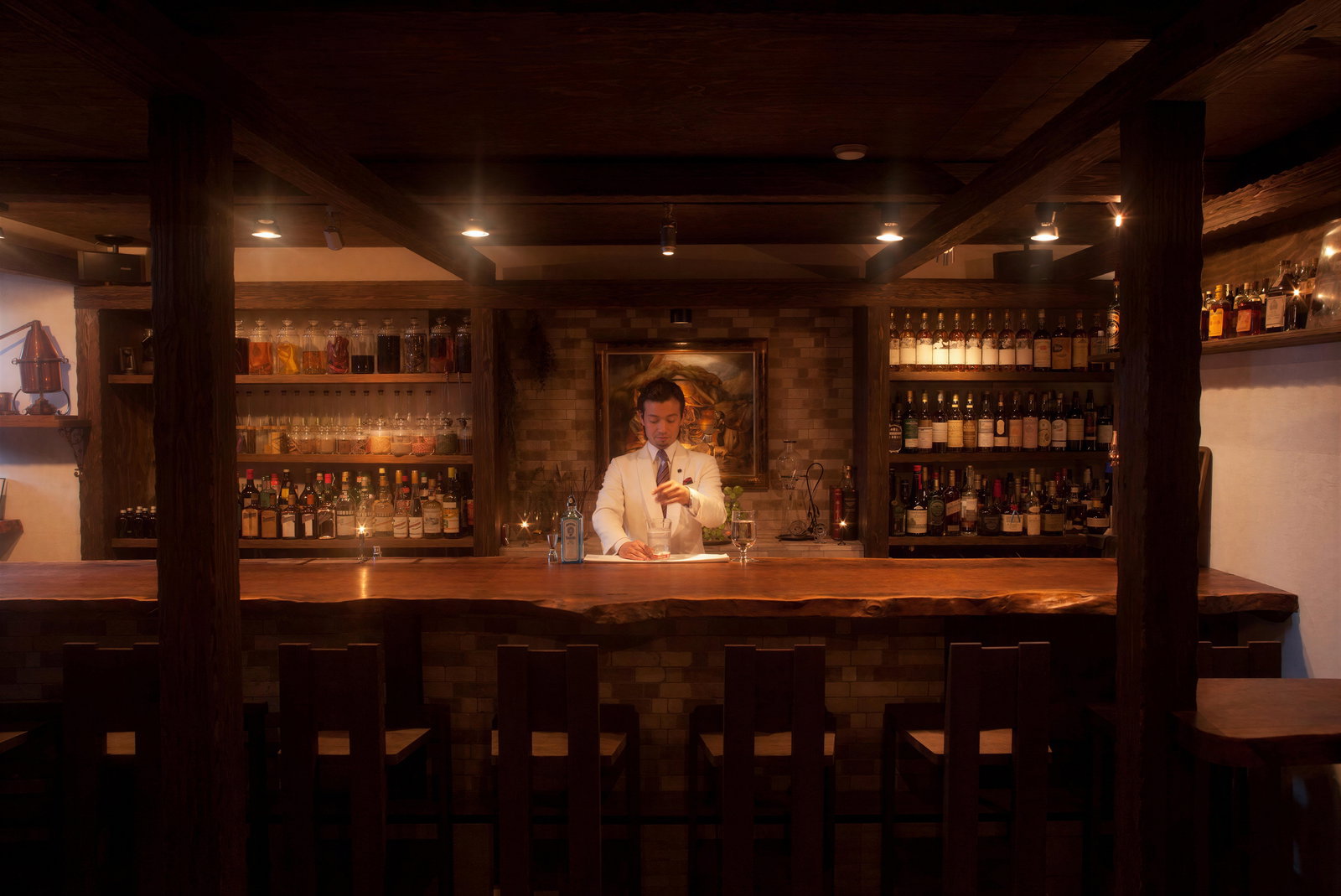 Bartender in a white suit mixes a drink at a dimly lit, wooden bar. Shelves of liquor bottles behind him create a warm, inviting atmosphere.