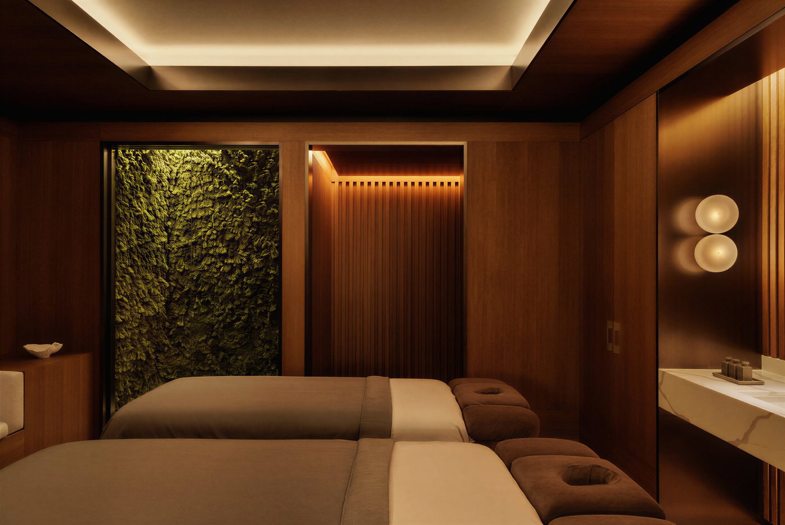 Auriga Spa Treatment Room at Capella Sydney with two massage tables, dim lighting, wooden walls, a green plant wall, and a modern sink with round lights, creating a tranquil ambiance.
