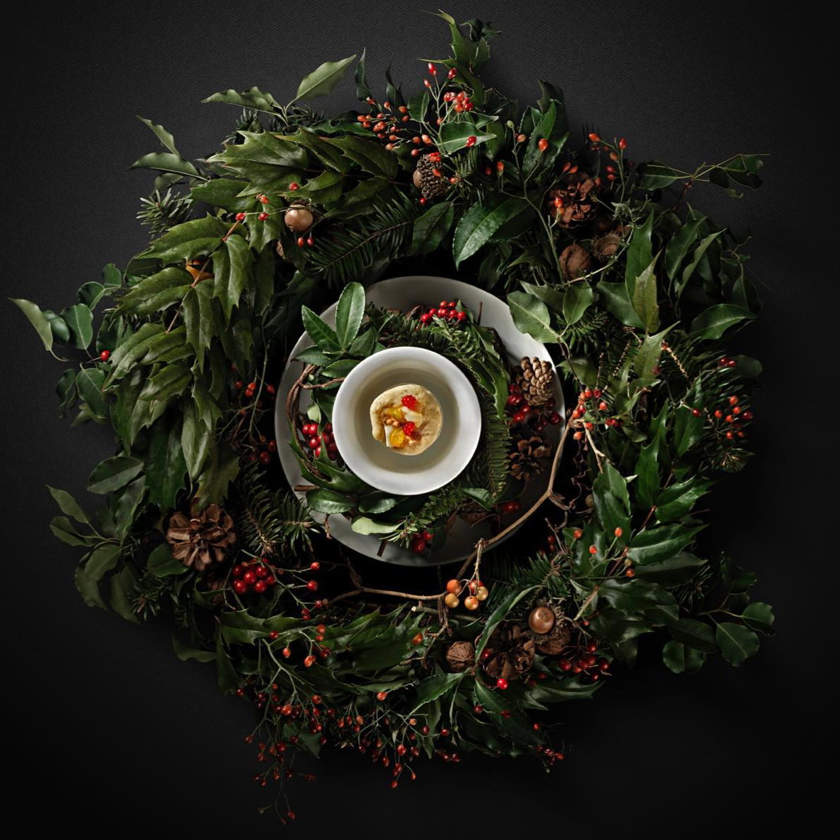 A white bowl with dessert sits atop a plate, surrounded by a wreath of green leaves, pinecones, and red berries on a dark background.