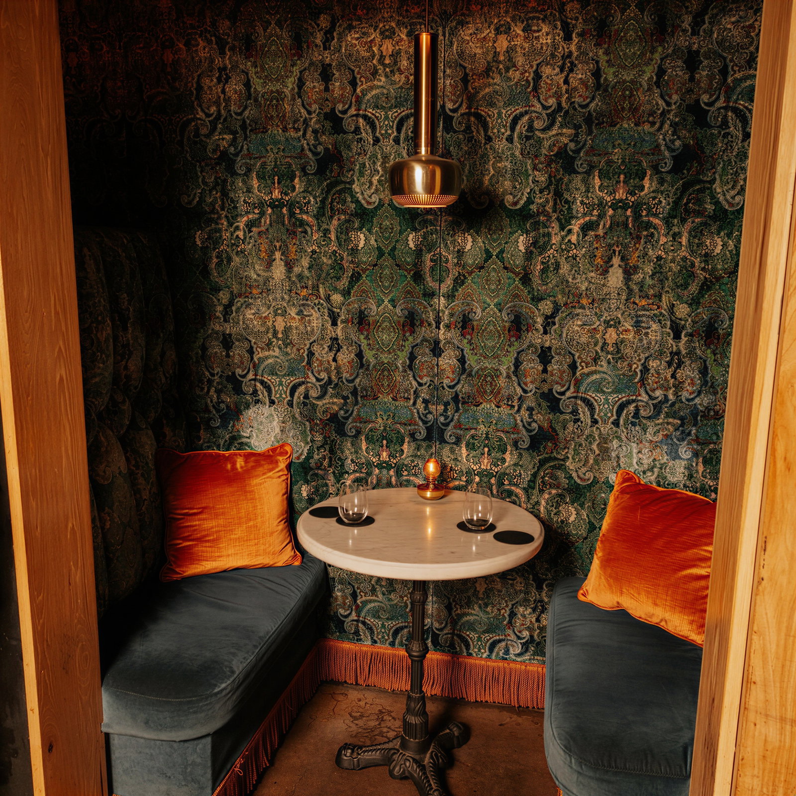 Cozy nook with velvet seats, orange pillows, ornate wallpaper, round table, and glasses of wine. Warm ambiance under a bronze pendant light.