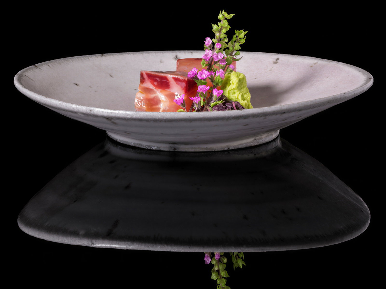 Gourmet dish on a white plate with slices of meat, green garnish, and pink flowers, set against a black background, reflecting below.