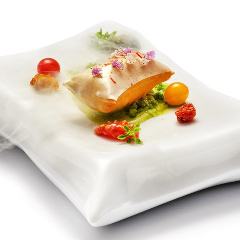 Elegant dish on a white pillow plate, featuring vibrant tomatoes, flowers, and herbs around a foam-topped item, with a misty effect.