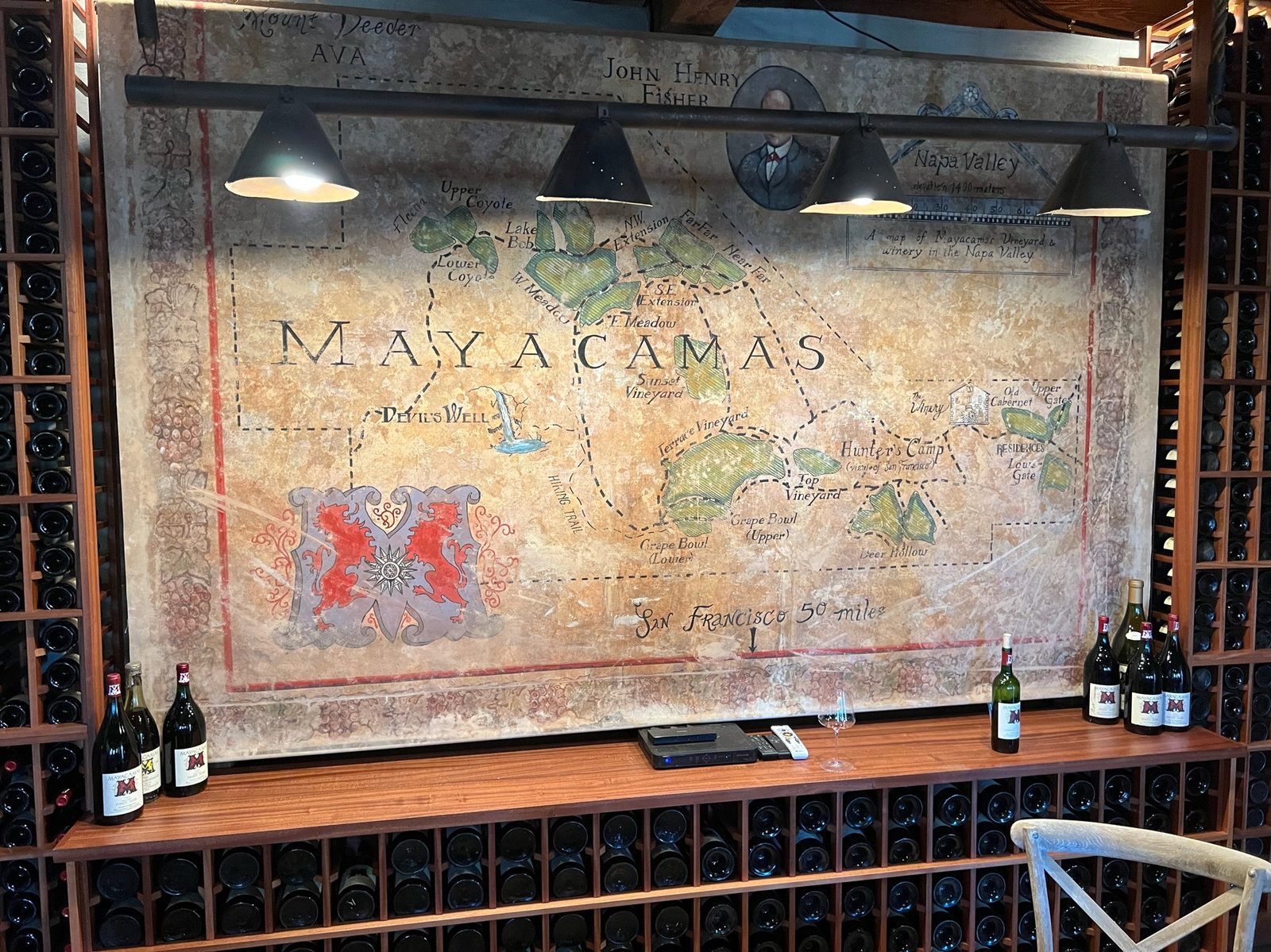 A large winery map labeled "MAYACAMAS" is displayed on a wall with a wooden shelf below, holding wine bottles. Wine racks fill the background.
