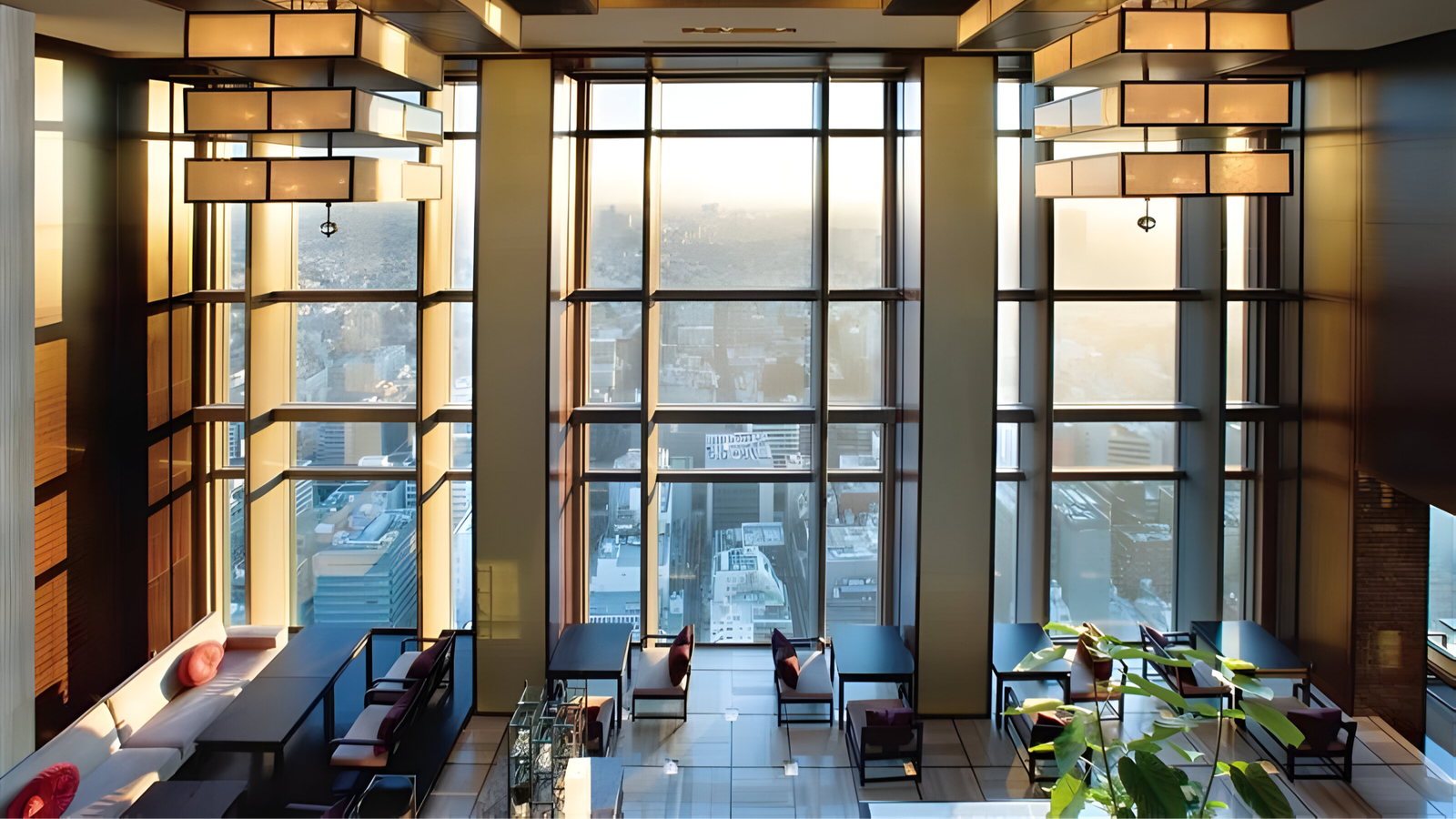 Modern restaurant interior with large windows overlooking a city skyline. Soft lighting, geometric chandeliers, and plush seating create a cozy ambiance.