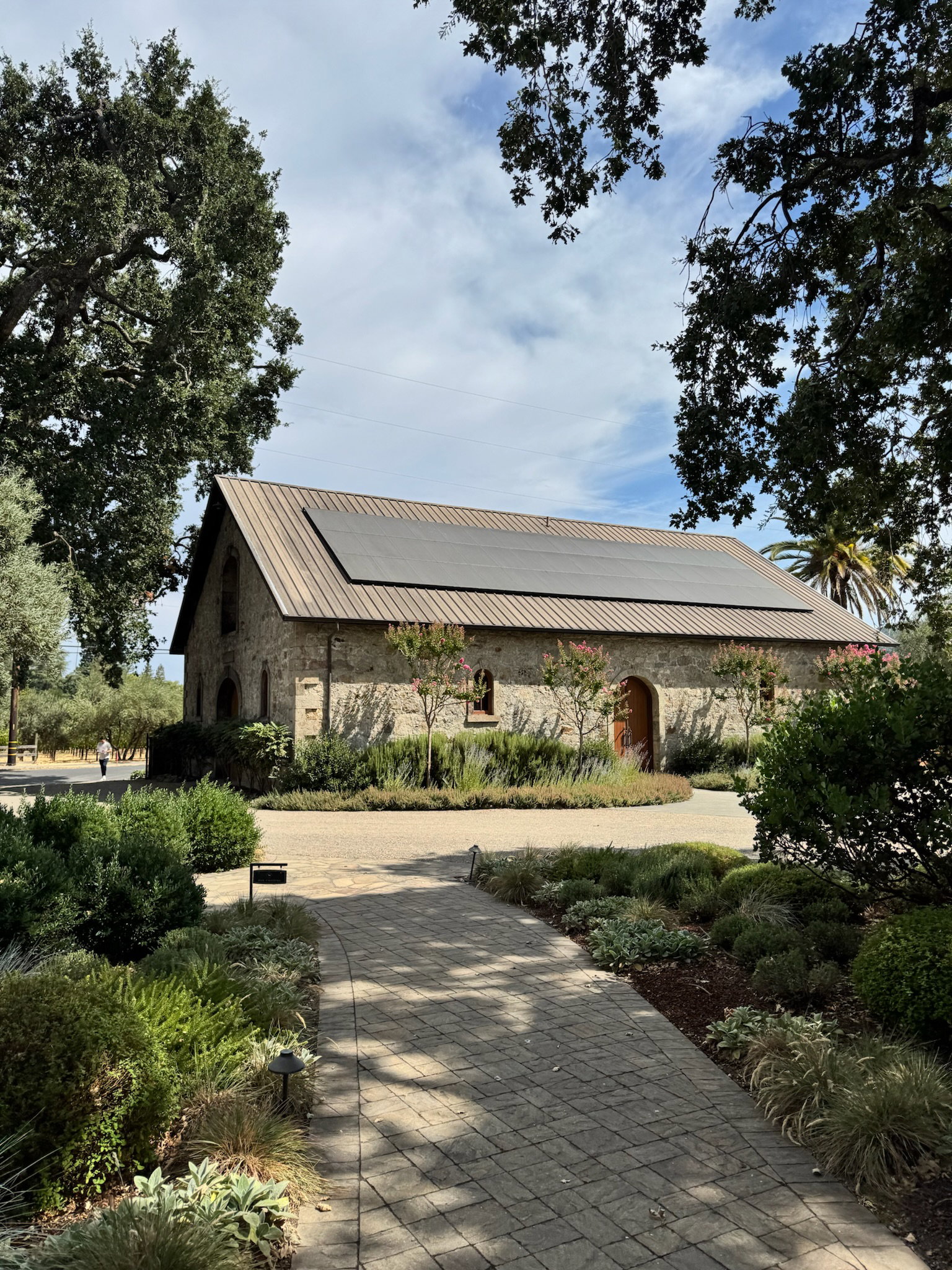 A stone building with a solar-paneled roof in a garden setting. Pathway leads to entrance, surrounded by lush greenery under a clear sky.