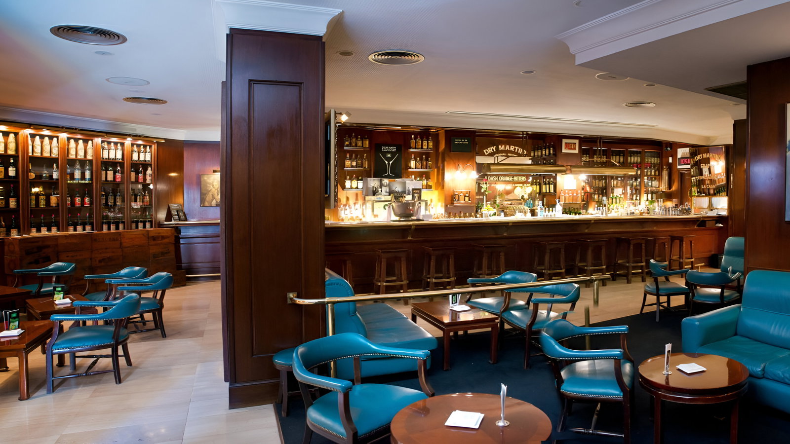 Empty bar with teal chairs and wooden tables, liquor bottles lined behind a long counter. Warm lighting creates a cozy atmosphere.