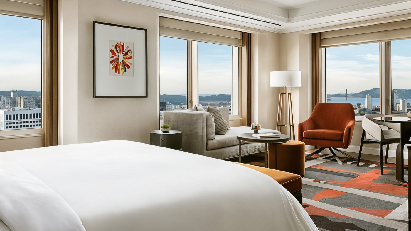 Modern hotel room with a city view, featuring a large bed, red-orange chair, and colorful abstract art. Neutral tones create a calm ambiance.
