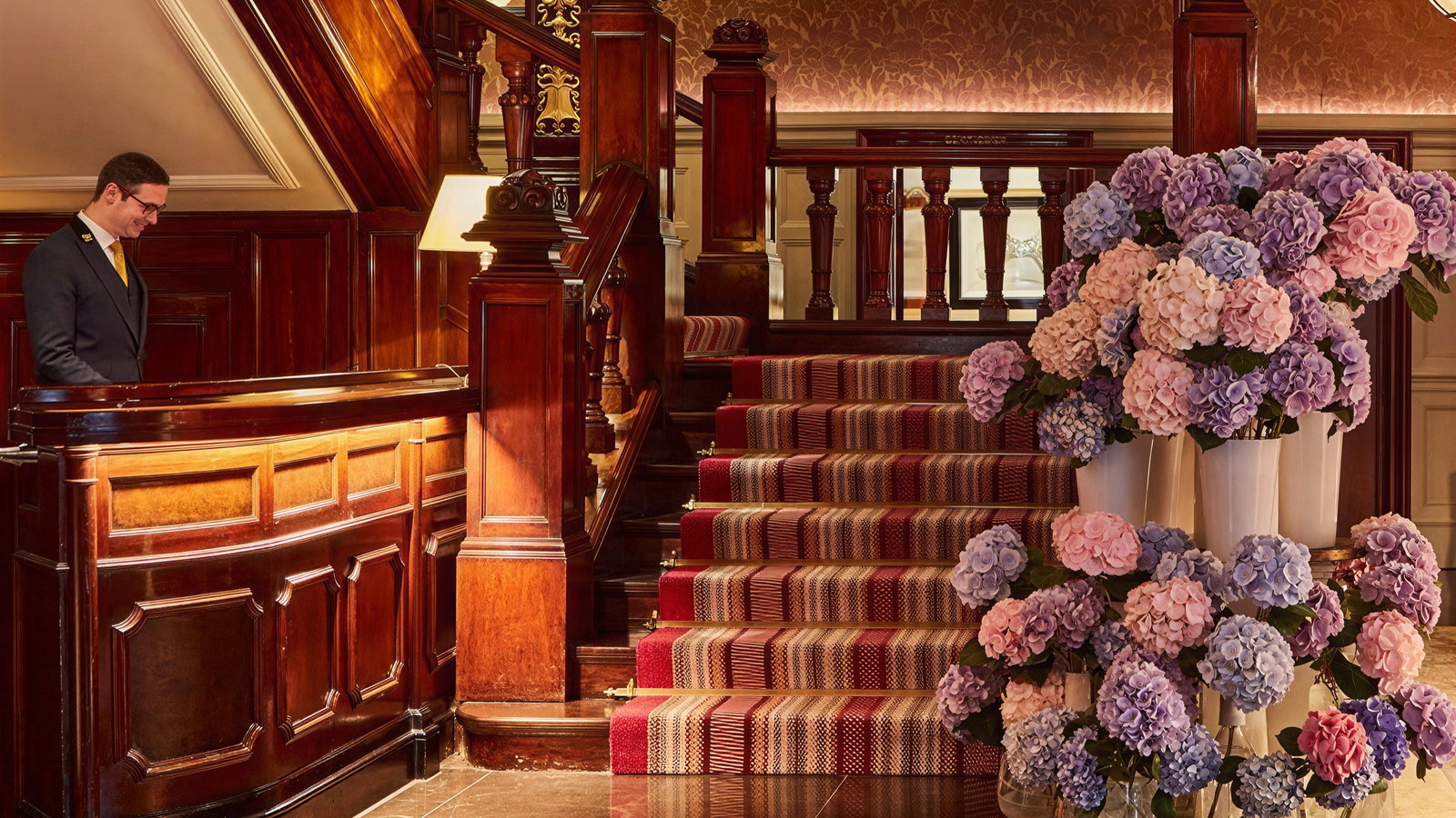 Elegant hotel lobby with a smiling doorman at a polished wooden desk. Red carpeted stairs and purple hydrangea bouquets create a refined ambiance.