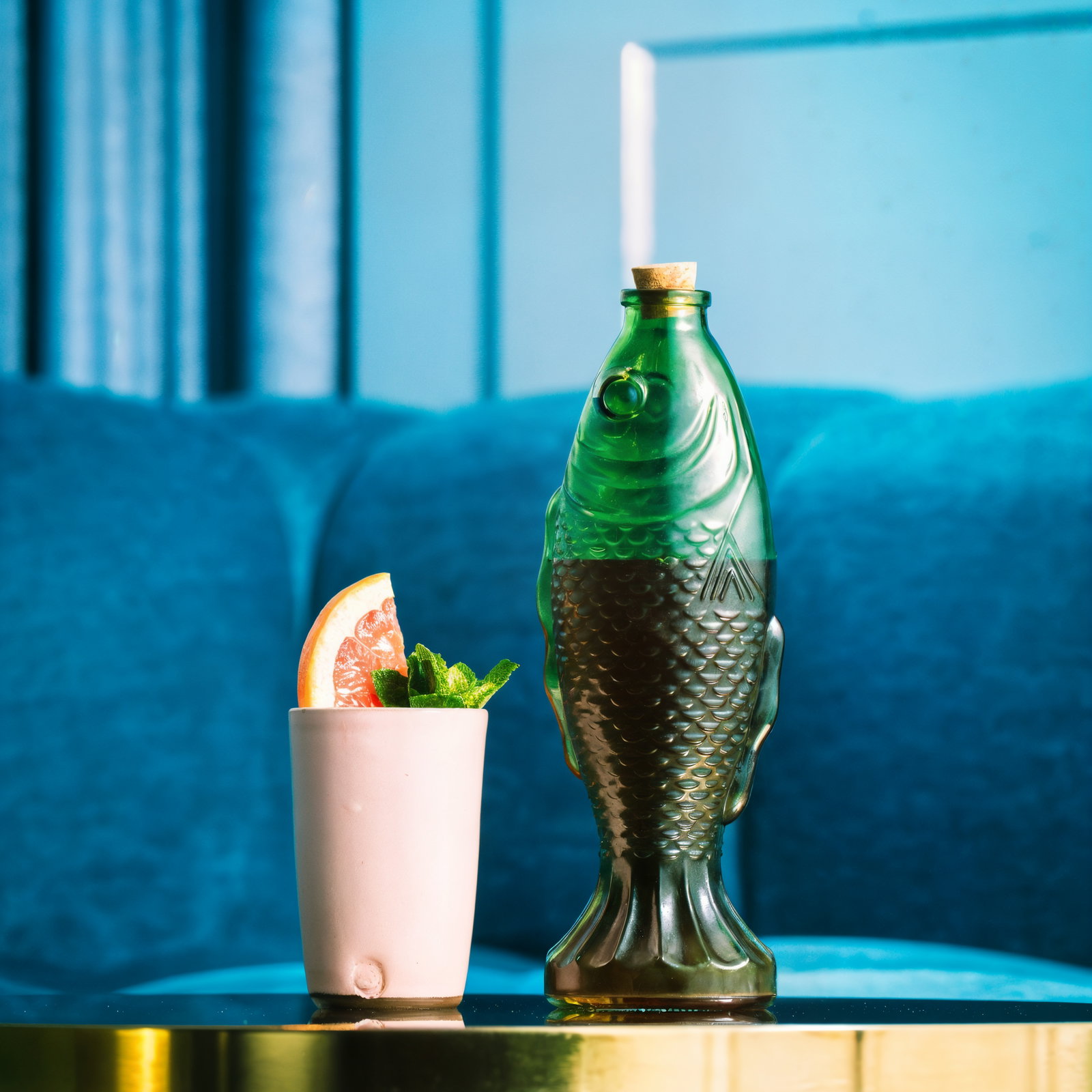 Green fish-shaped bottle and a pink cup with grapefruit and mint on a gold table. Blue sofa in the background.