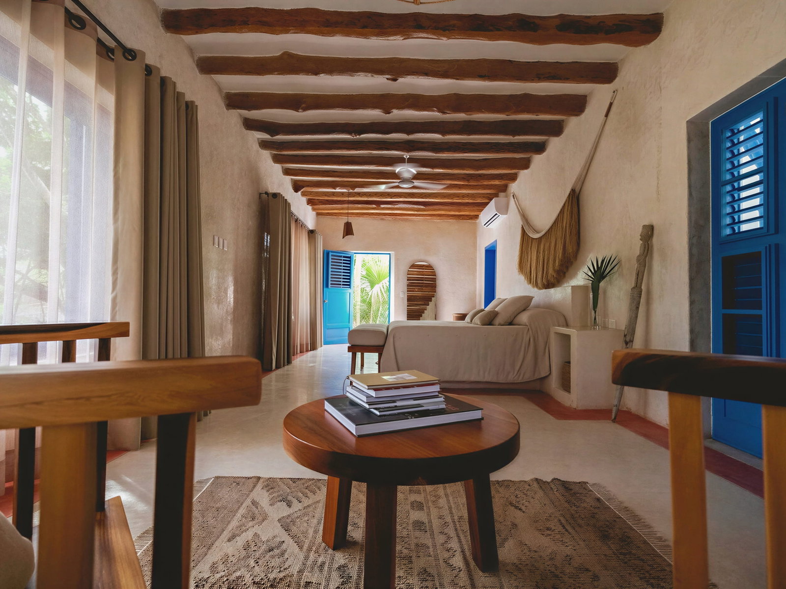 Cozy Casa de Piedra room at Wakax Hacienda with beige walls, wooden beams, blue doors, and a bed. A table with books and a rug in the foreground; tropical view outside.