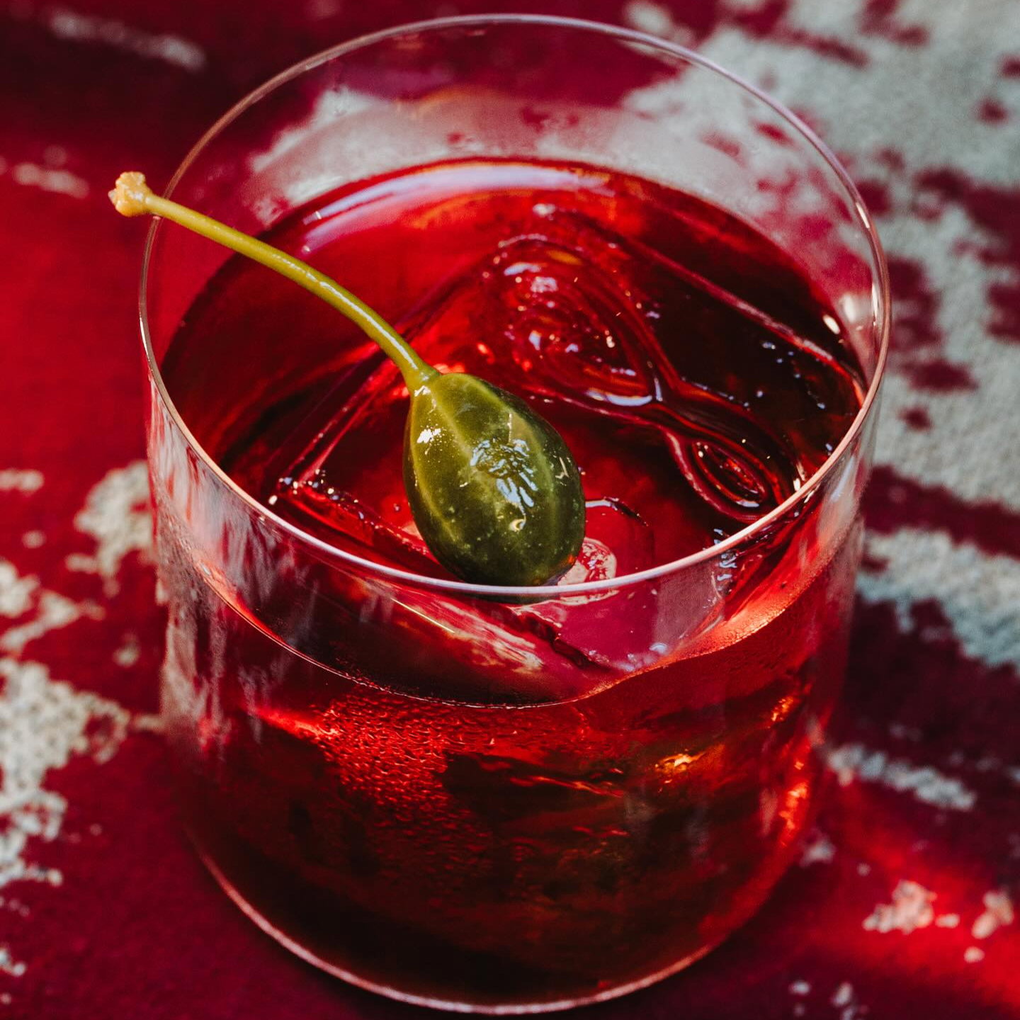 Glass of red cocktail with ice cube and caper garnish on a red and white textured surface. Vibrant and refreshing mood.