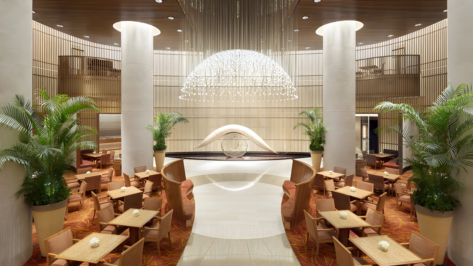 Elegant hotel lobby with a textured ceiling, large pillars, potted plants, and wooden furniture on a patterned red carpet. Calm ambiance.