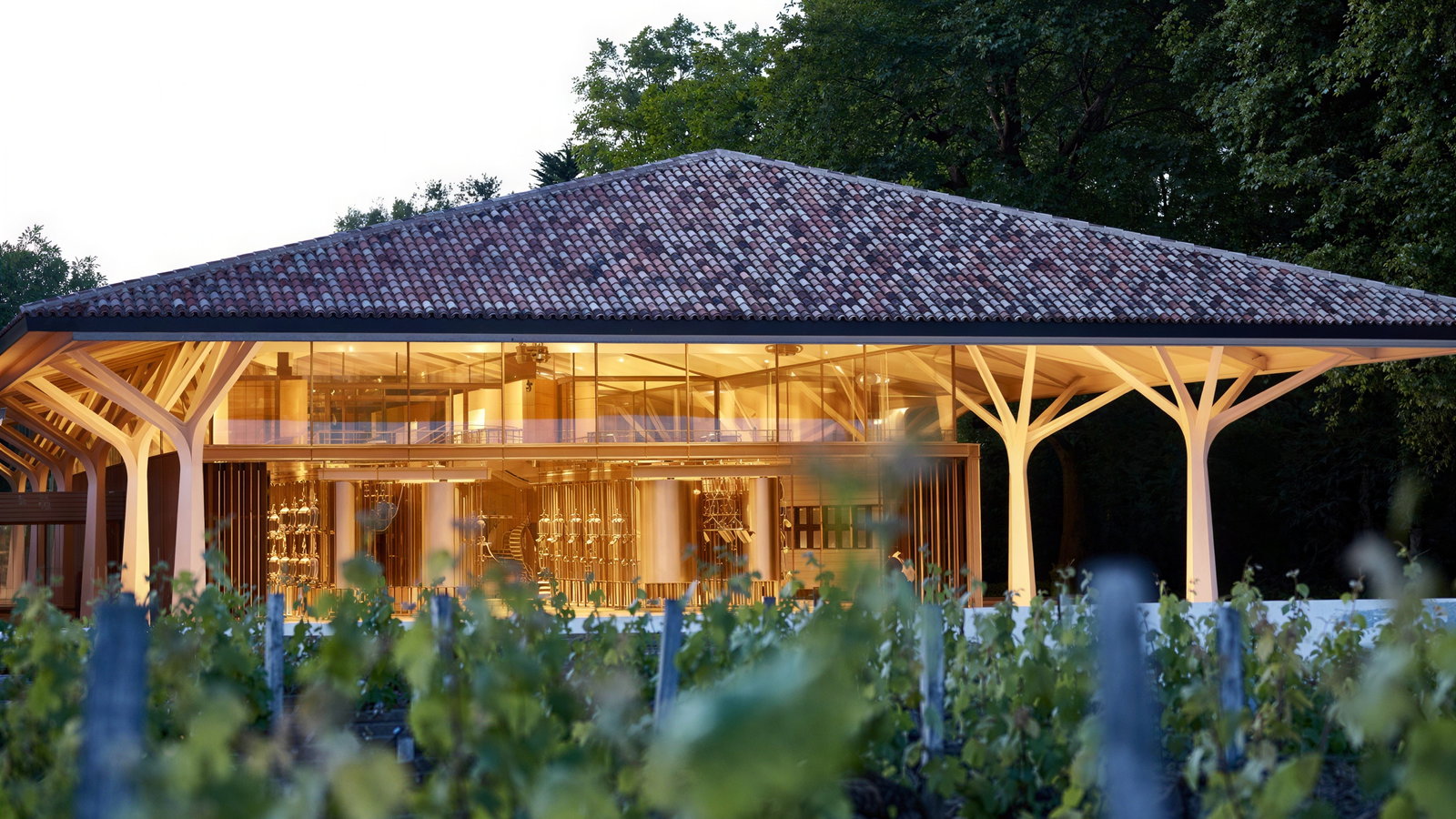 Modern glass building with a red-tiled roof amidst lush greenery. Warm interior lighting visible, creating a welcoming ambiance.